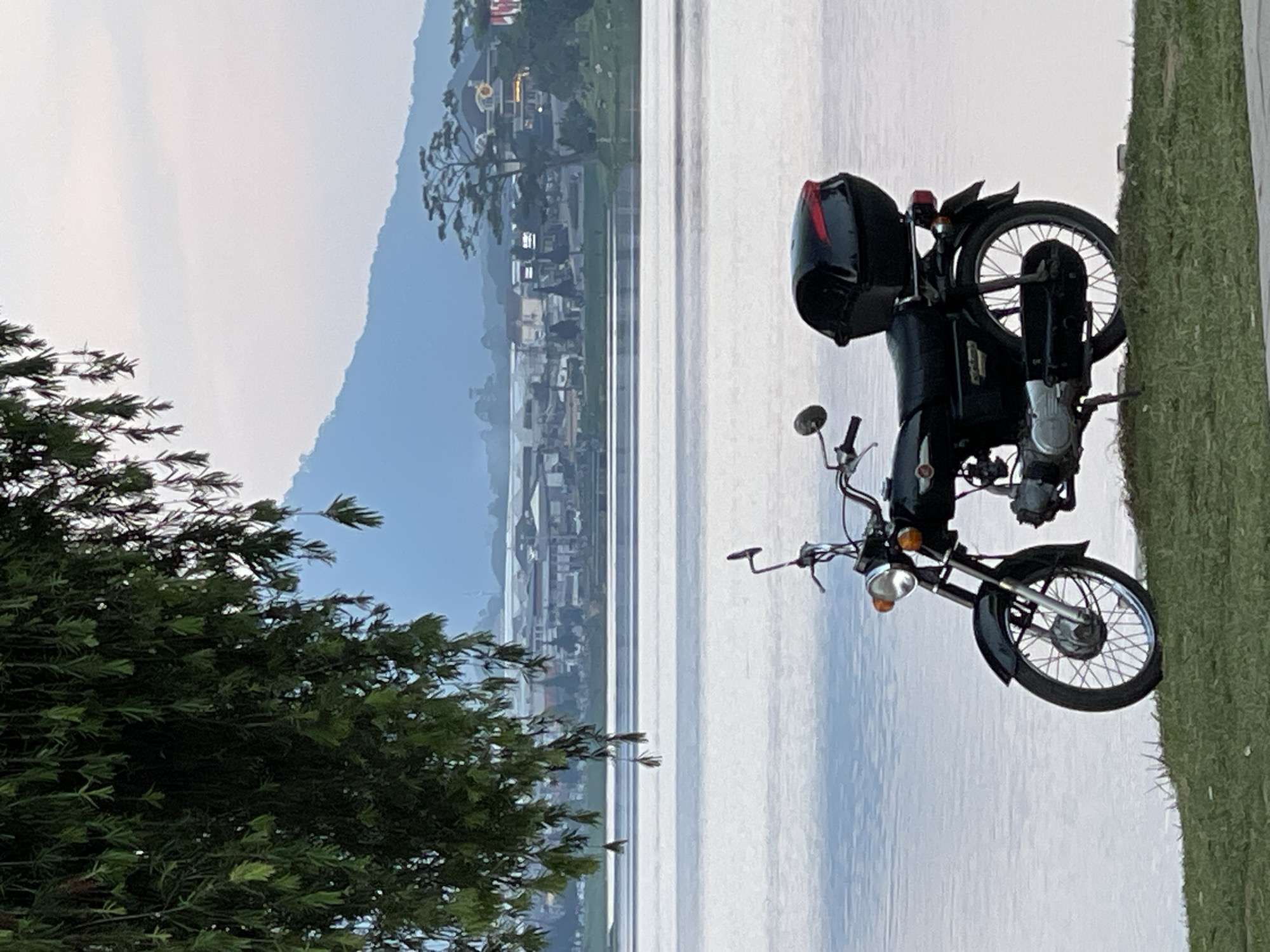 A black motorcycle with a black helmet strapped to its rear is parked on a grassy strip along a body of water. In the background, buildings and mountains are visible under a light sky. Trees are present in the foreground. The scene suggests an outdoor setting in a location near a city or town situated by a lake or reservoir, possibly in Da Lat, Vietnam.