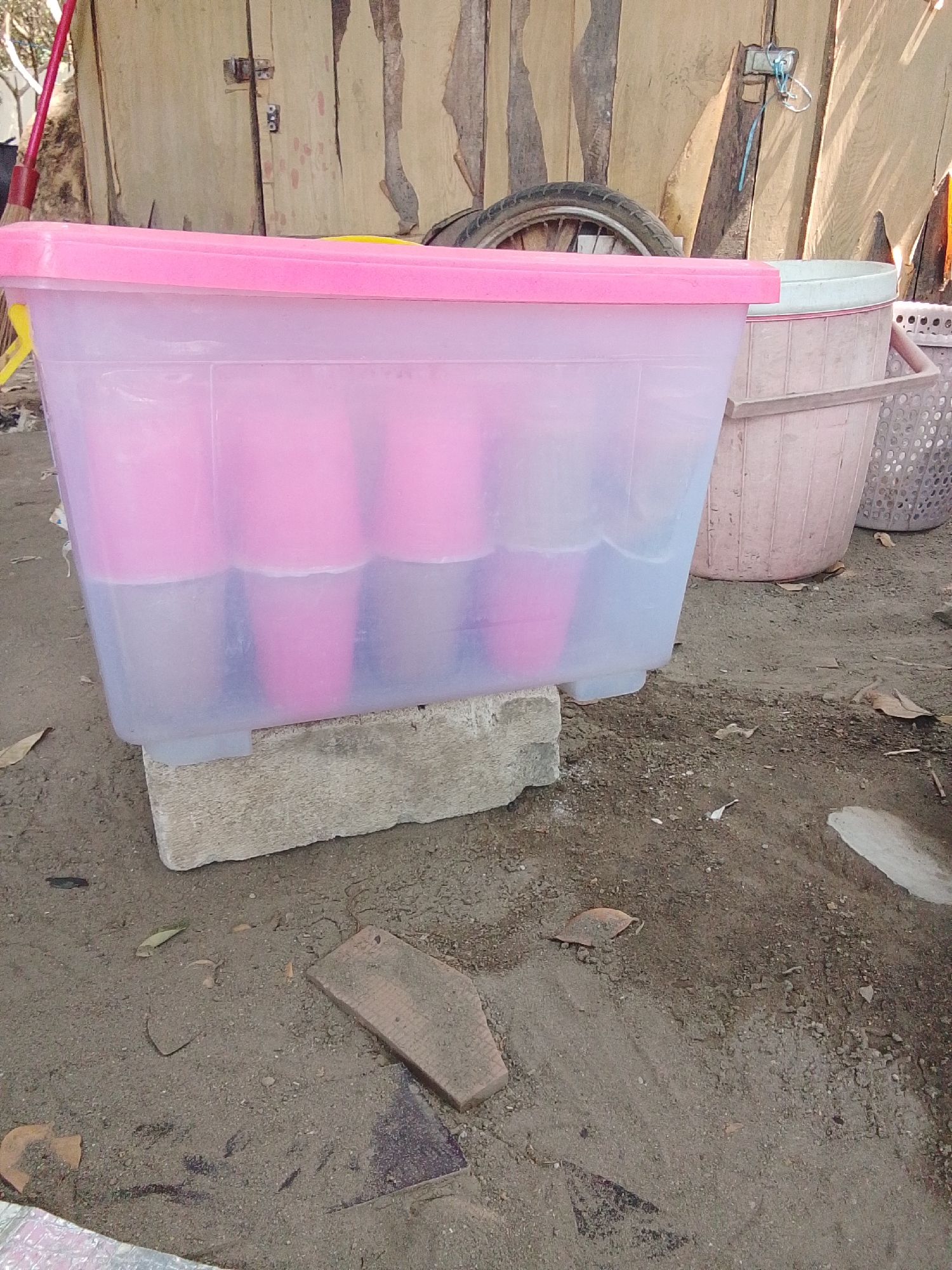 An outdoor scene in Jogoroto, Indonesia, captures a clear plastic storage bin with a pink lid, filled with water and pink cylindrical objects, likely cups. The bin rests on a light gray concrete block, which is placed on a dirt ground scattered with small stones and dried leaves. Behind the bin, a portion of a wooden wall is visible, with a metal latch and blue string attached. To the right of the plastic bin, a larger, light pink plastic tub is present, and further to the right, a woven basket is partially visible. In the background, a bicycle wheel and tire are visible leaning against the wooden wall. A red broom with a yellow handle stands to the left of the scene. The ground is uneven and appears to be an outdoor area with some debris.