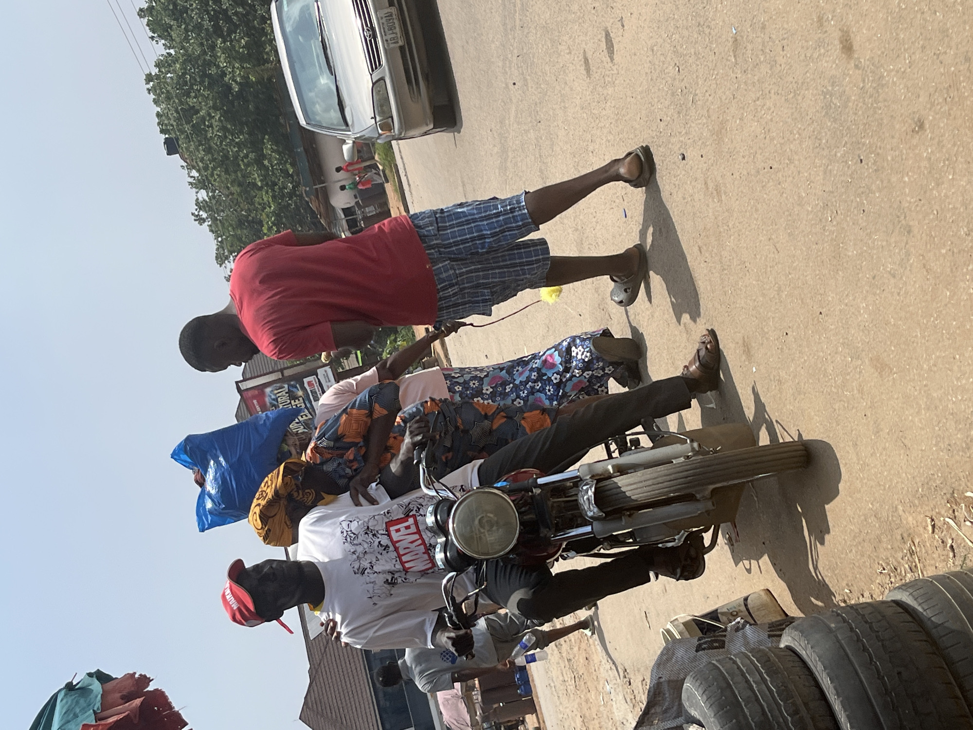 The image depicts a street scene in Abuja, Nigeria. The primary subjects are several people and a motorcycle, suggesting a mode of transportation or a vendor's setup. One person in a red shirt and blue plaid shorts stands holding the handlebars of the motorcycle, appearing to be either preparing to ride or having just stopped. Another person, wearing a pink and blue patterned dress, is seated on the back of the motorcycle, with their arm resting on the shoulder of the person in front. A third person in a white t-shirt with a Marvel logo and a red cap is also on or near the motorcycle, holding a blue bag.
The setting is a paved road, with a silver car visible in the background. Trees and some buildings are also present, hinting at an urban or semi-urban environment. The lighting suggests it is daytime, likely late morning or afternoon, with a clear sky. The overall mood seems casual, with people gathered around the motorcycle.
There is visible text on the white t-shirt, which reads "MARVEL". Another small text is visible on a sign in the background. The presence of stacked tires in the foreground might indicate a tire shop or a place where tires are sold or repaired. The image is oriented such that the scene appears rotated.