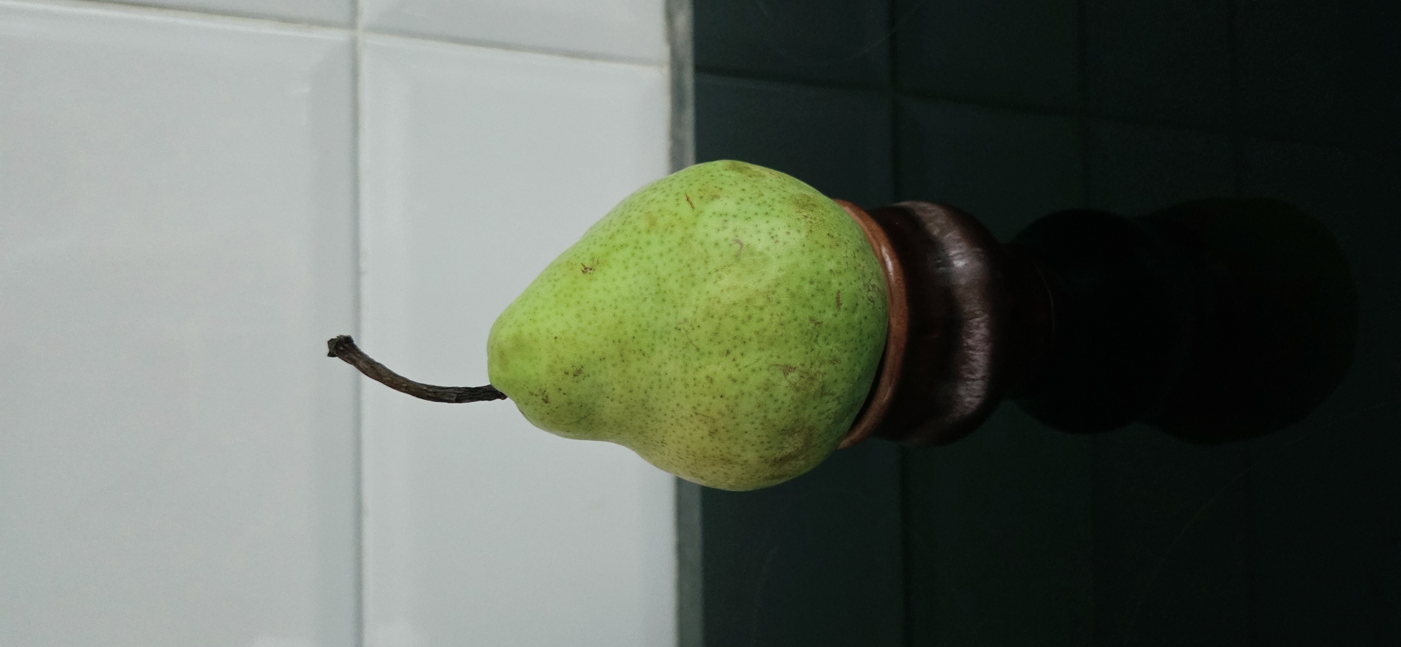 A single green pear with a dark stem sits atop a small, dark, rounded pedestal. The pear is positioned in the center of the frame, with its wider base resting on the pedestal. The background consists of a white tiled wall, with a dark, reflective surface in the foreground. The image is a still life, with no people or animals present. The lighting is even, creating a soft shadow of the pear and pedestal on the reflective surface. The scene is clean and minimalist. There is no information available about the location in Lipetsk, Russia.