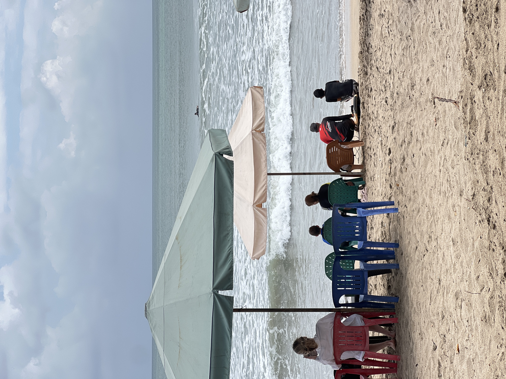The image depicts a beach scene in Kuta, Indonesia, under a cloudy sky. In the foreground, sand covers the ground, with a few scattered twigs. Two large beach umbrellas, one green and one cream-colored, provide shade. Several plastic chairs are arranged on the sand.
Under the green umbrella, a woman in a white shirt is seated in a red chair, looking at her phone. To her right, a young person in a blue shirt sits in a green chair, and another person in a black shirt is in a blue chair.
Under the cream-colored umbrella, a person in a red and black shirt is sitting on a brown chair, facing the ocean. Beside them, a person in a black shirt is sitting on the sand, with a surfboard next to them.
The ocean in the background has moderate waves with white foam. A lone surfer is visible in the distance, paddling on a surfboard. The water appears slightly choppy. The overall atmosphere suggests a relaxed day at the beach.