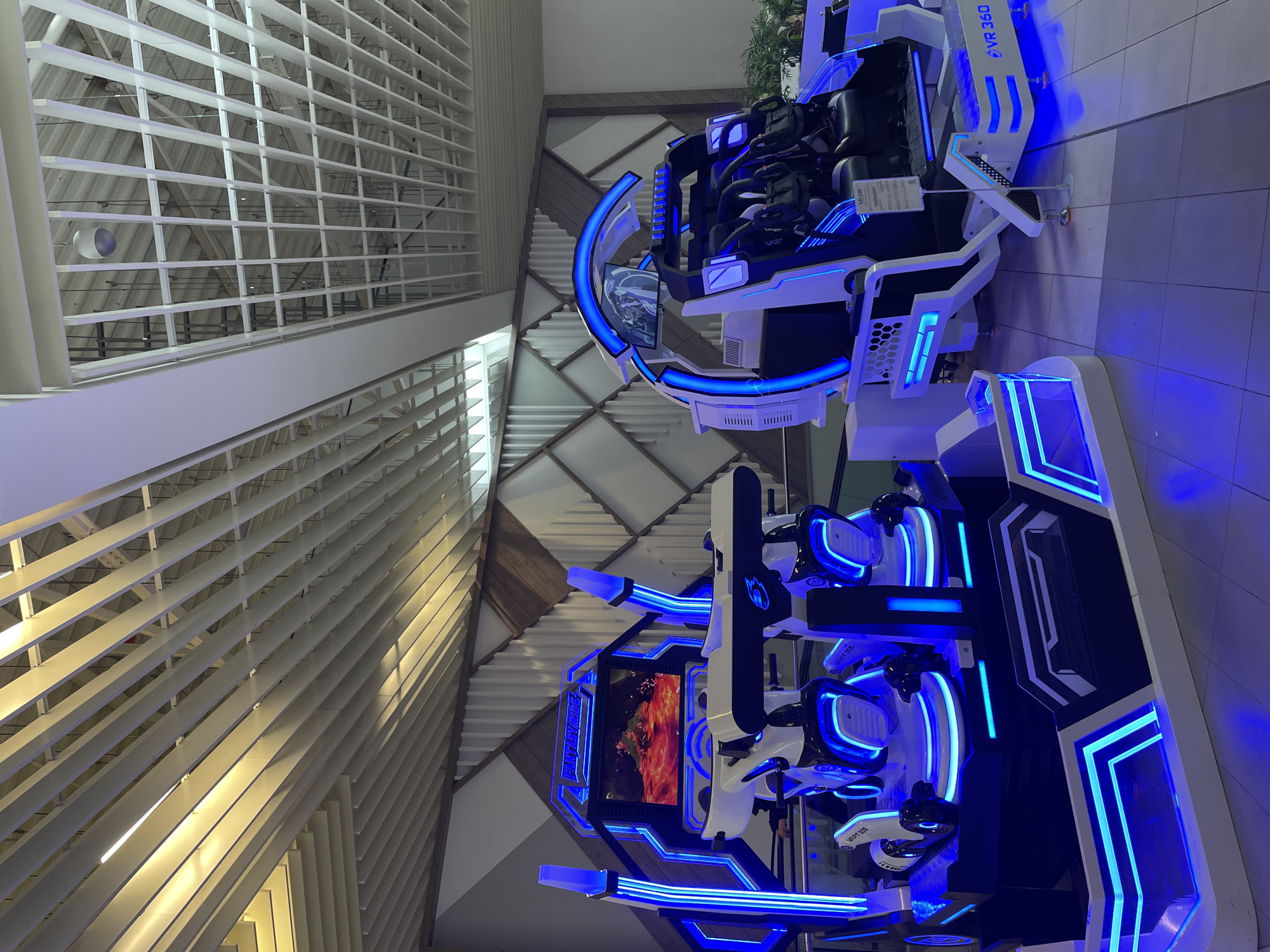 The image depicts the interior of a building in Kazan, Russia, showcasing two virtual reality (VR) simulator machines with white and black frames illuminated by blue LED lights. The machines, labeled "VR 360" and "FANTASYRIDE VRT 005," are designed to seat multiple people. One features a display screen showing a volcanic scene. The backdrop includes a white, geometrically patterned ceiling structure with visible internal support beams. The lighting conditions suggest artificial illumination.