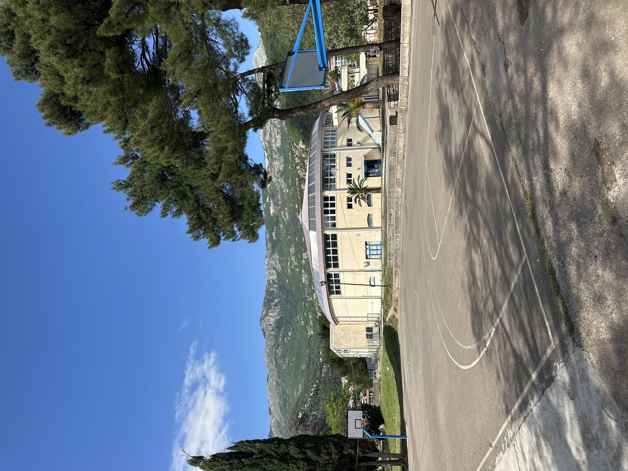 The image shows a basketball court adjacent to a building, with mountains in the background and a clear blue sky. The building is light yellow with a curved roof and multiple windows. There are two basketball hoops visible on the court. Trees surround the area, and a palm tree is near the building. The location is likely in or near the city of Budva, Montenegro, due to the presence of the mountains and coastal landscape characteristic of the region. No people are visible in the image.
