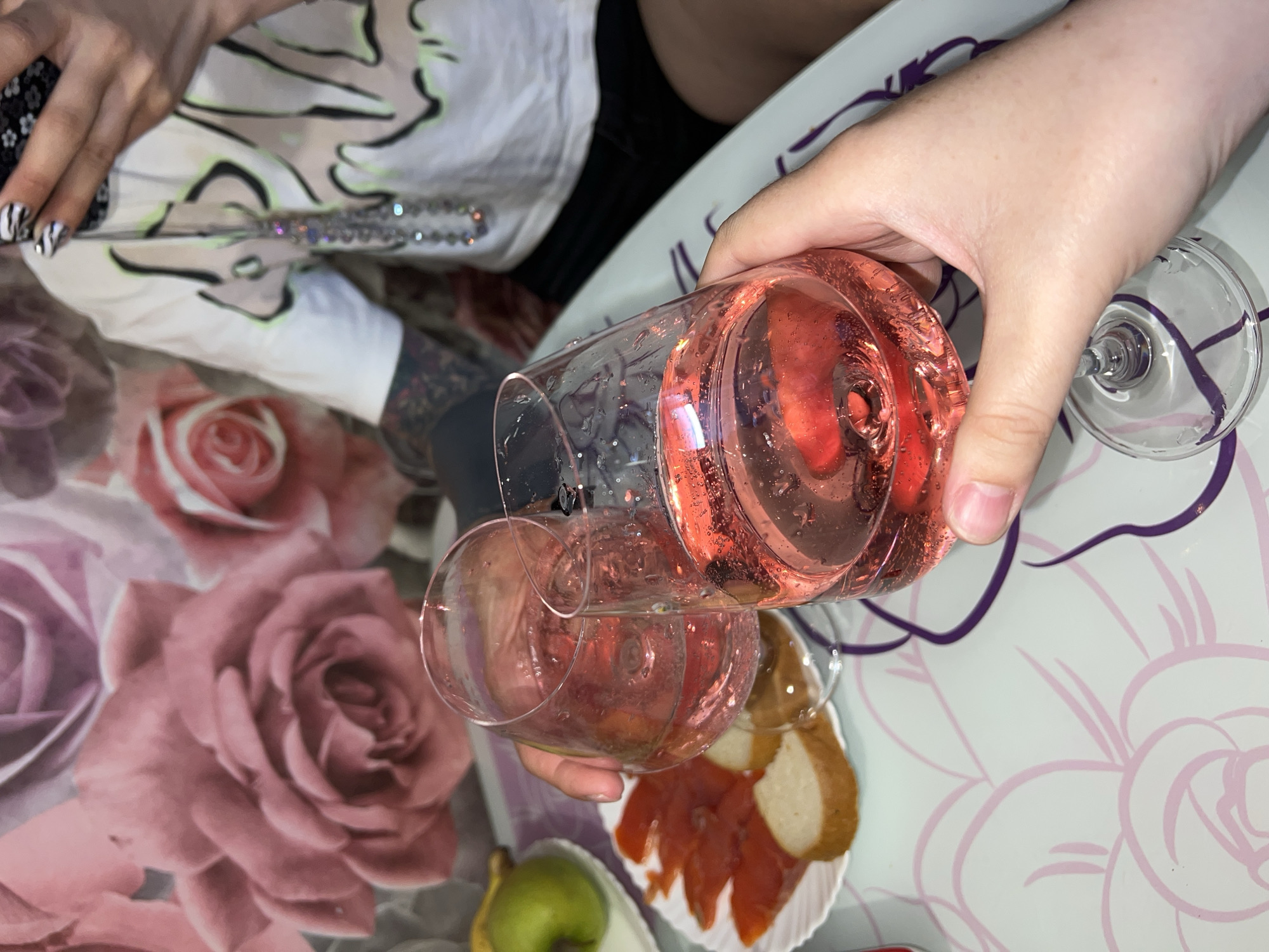 A high-angle shot shows a person pouring a pink liquid into a clear wine glass. The liquid comes from another clear wine glass. Next to the receiving glass is a plate with what appear to be slices of salmon and bread. A green pear sits near the plate. The setting is an indoor table with a floral patterned surface. A person wearing a white shirt with black sleeves sits to the left. This person has black and white patterned nails and is holding a bedazzled object. The location is Noviy, Russia.