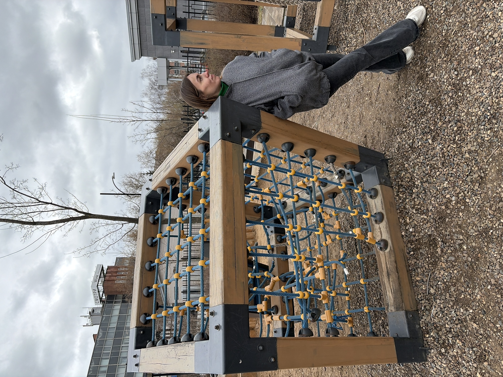 This image, taken in Izhevsk, Russia, depicts an outdoor scene at a playground or park on an overcast day. A young woman is the central subject, sitting on a sturdy, wooden climbing structure. She has light brown hair and is wearing a grey, possibly felted, long-sleeved top over a green turtleneck, paired with dark grey or black wide-leg jeans and white sneakers. Her posture is relaxed, and she looks to her right with a neutral, possibly contemplative, expression.
The prominent object is the cubic climbing structure, constructed from light-colored wooden beams connected by dark grey or black metal brackets. Inside this frame, a dense blue rope net forms a climbing grid, featuring numerous yellow and black plastic connectors. The ground beneath and around the structure is covered in light-colored gravel.
In the background, bare deciduous trees suggest either late winter or early spring. Beyond the trees, several buildings are visible, including a modern structure with a grid of windows and another older, reddish-brown building. On top of the older building, a sign clearly displays the text "СМОТРИ Wink," which translates to "WATCH Wink," indicating a streaming service advertisement. The sky is uniformly cloudy and grey, suggesting a cool, possibly dull, daytime atmosphere.