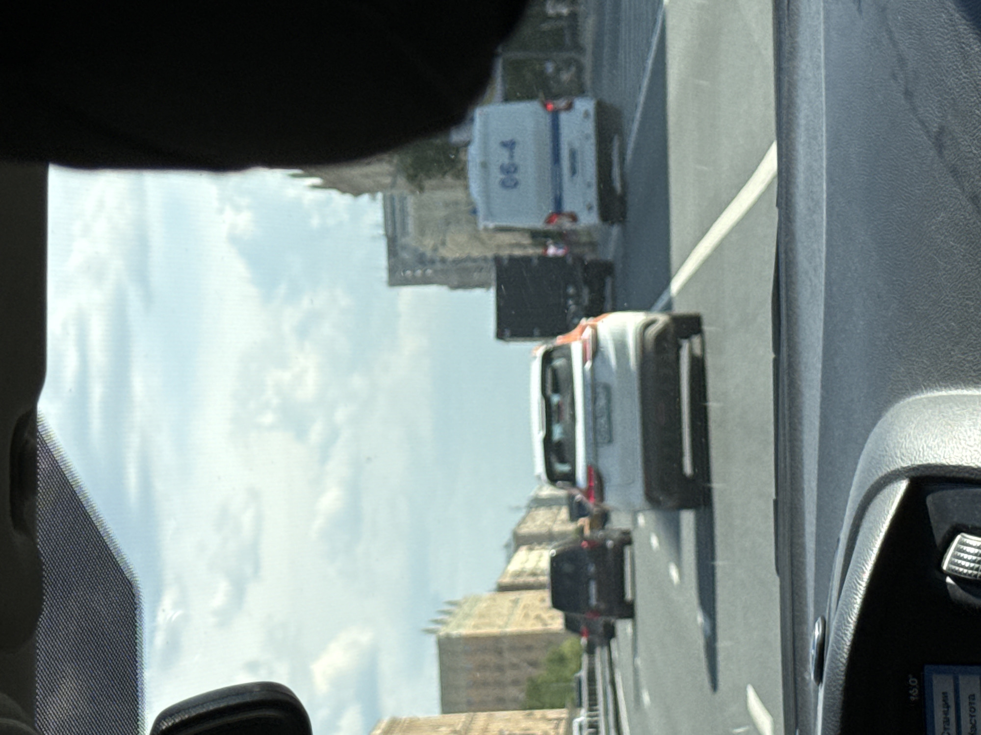 The image is taken from inside a vehicle, looking through the front windshield at a city street on a bright, partly cloudy day. The perspective is looking forward, with parts of the car's interior visible in the foreground.
In the immediate foreground, sections of a dark dashboard, a side pillar, and what appears to be a rearview mirror assembly are present. A digital display on the dashboard, seen rotated, shows Cyrillic text including "станция" (station) and "высота" (height), with the number "16.0" next to "высота".
The scene outside the vehicle features a multi-lane asphalt road with white lane markings. Several vehicles are ahead, suggesting traffic or movement. A white sedan is partially visible in the near-middle ground. Further down the road, a prominent white utility or service van displays the numbers "06-4" on its rear. Behind this van, a dark sedan is visible, followed by other indistinct vehicles in the distance.
The background is filled with multi-story buildings featuring classical or traditional architectural styles, including visible rooflines and intricate facade details, consistent with historical urban centers. The sky above is bright with scattered clouds. No people are clearly visible in the image. The overall impression is of driving or being stopped in traffic within a bustling city environment.