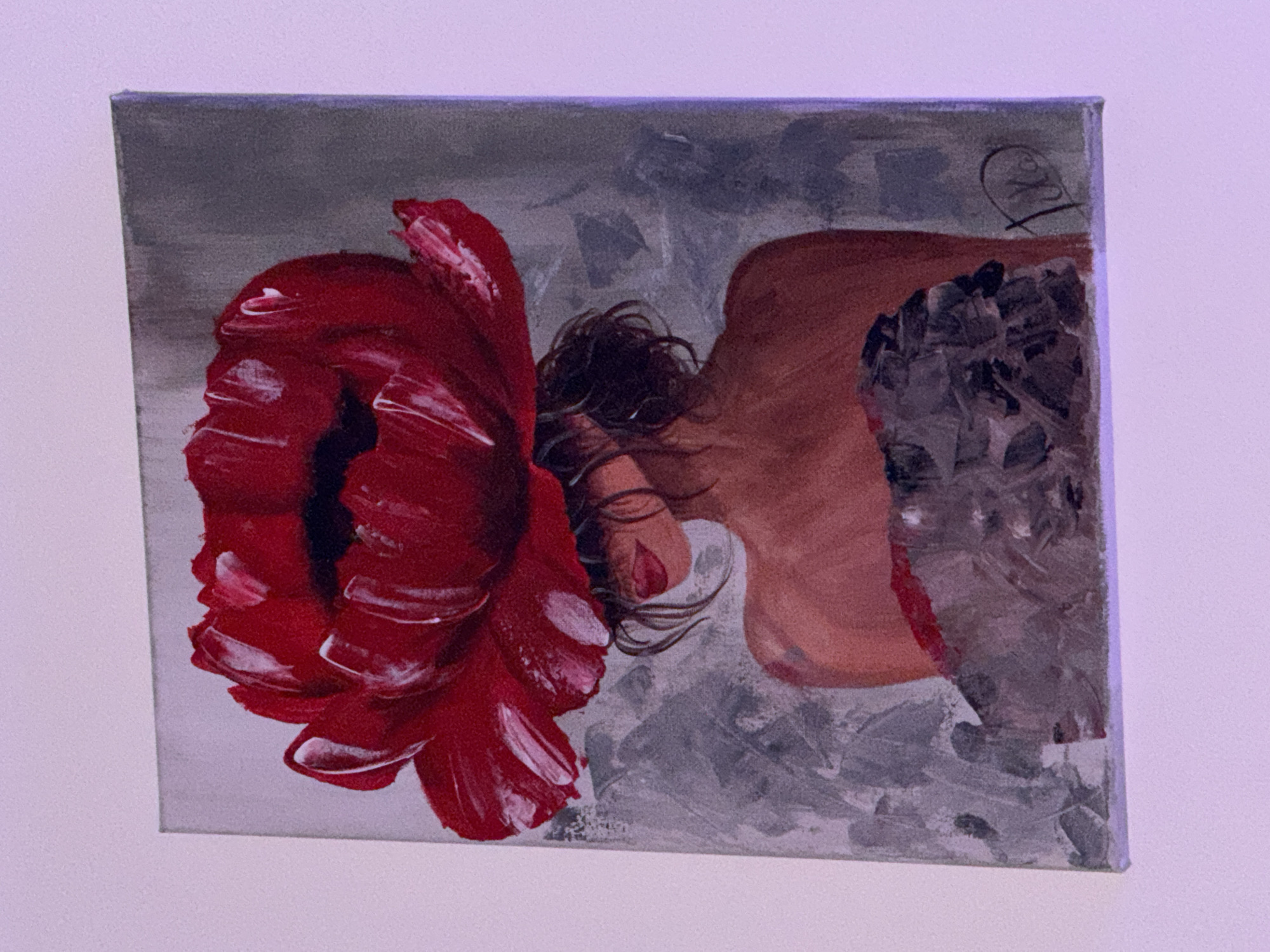 The image shows a painting of a woman with a large red flower as her head. The painting is rendered in acrylics on canvas and features a muted grey background with textured brushstrokes. The woman's skin tone is a warm brown, and her lips are painted a vibrant red. Her hair is dark and styled loosely around her face, which is mostly obscured by the flower. She is depicted from the shoulders up, with a portion of a dark, textured garment visible at the bottom. A signature, appearing to be "Tiko," is visible in the bottom right corner of the painting. The painting is hanging on a light-colored wall, with a purple light source casting a subtle glow on the scene. There is no discernible location in Plovdiv, Bulgaria, or any specific environmental context provided by the image.