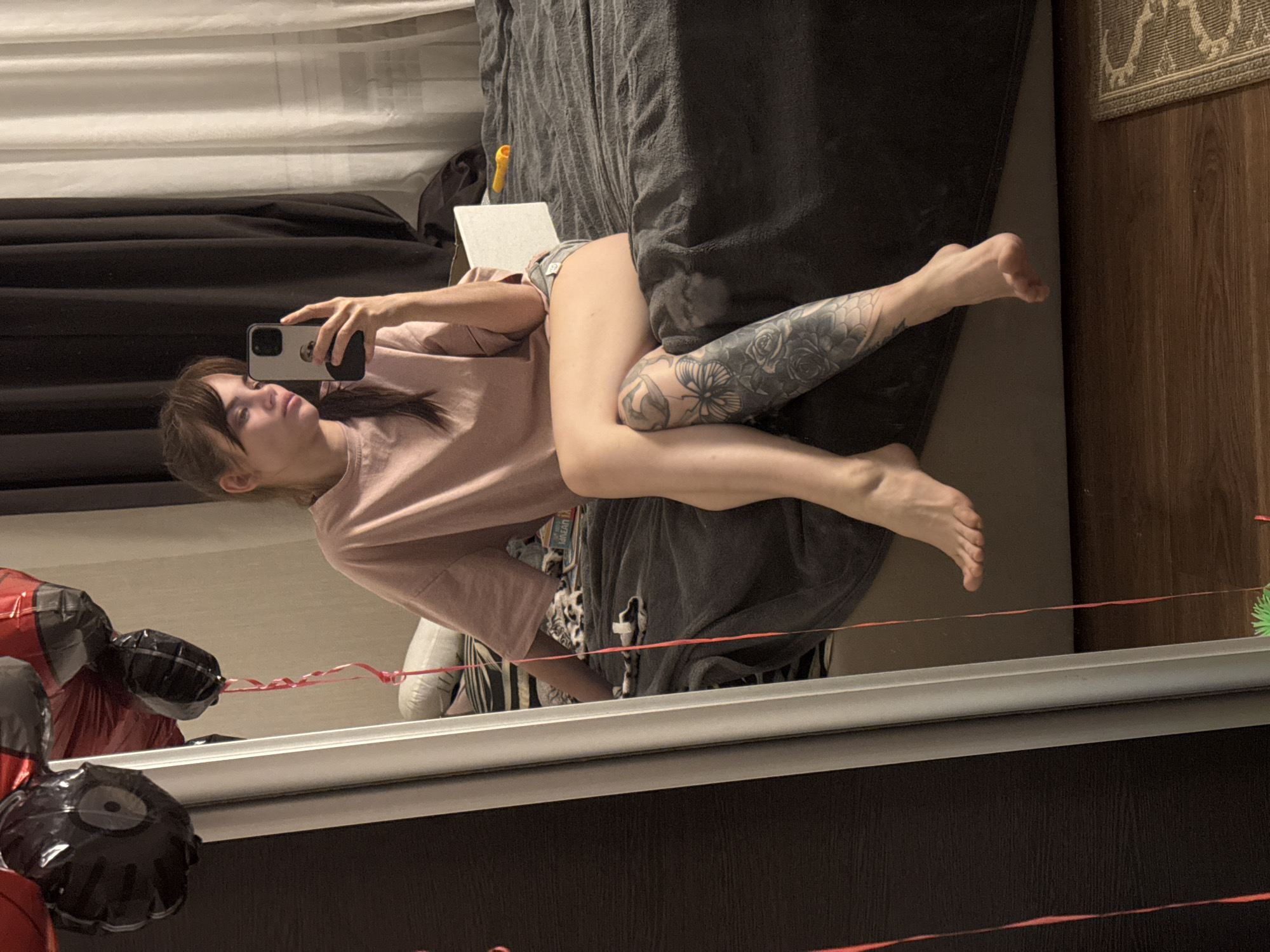 A woman is taking a selfie in a mirror. She is lying on her side on a bed, with her legs outstretched and tattooed. She is wearing a pink t-shirt and shorts. A phone is in her left hand. The bed has a gray comforter. The room has a wooden floor and white curtains. Balloons are visible in the mirror reflection. The location is likely indoors. There is no indication of the city of Rostov-on-Don, Russia.