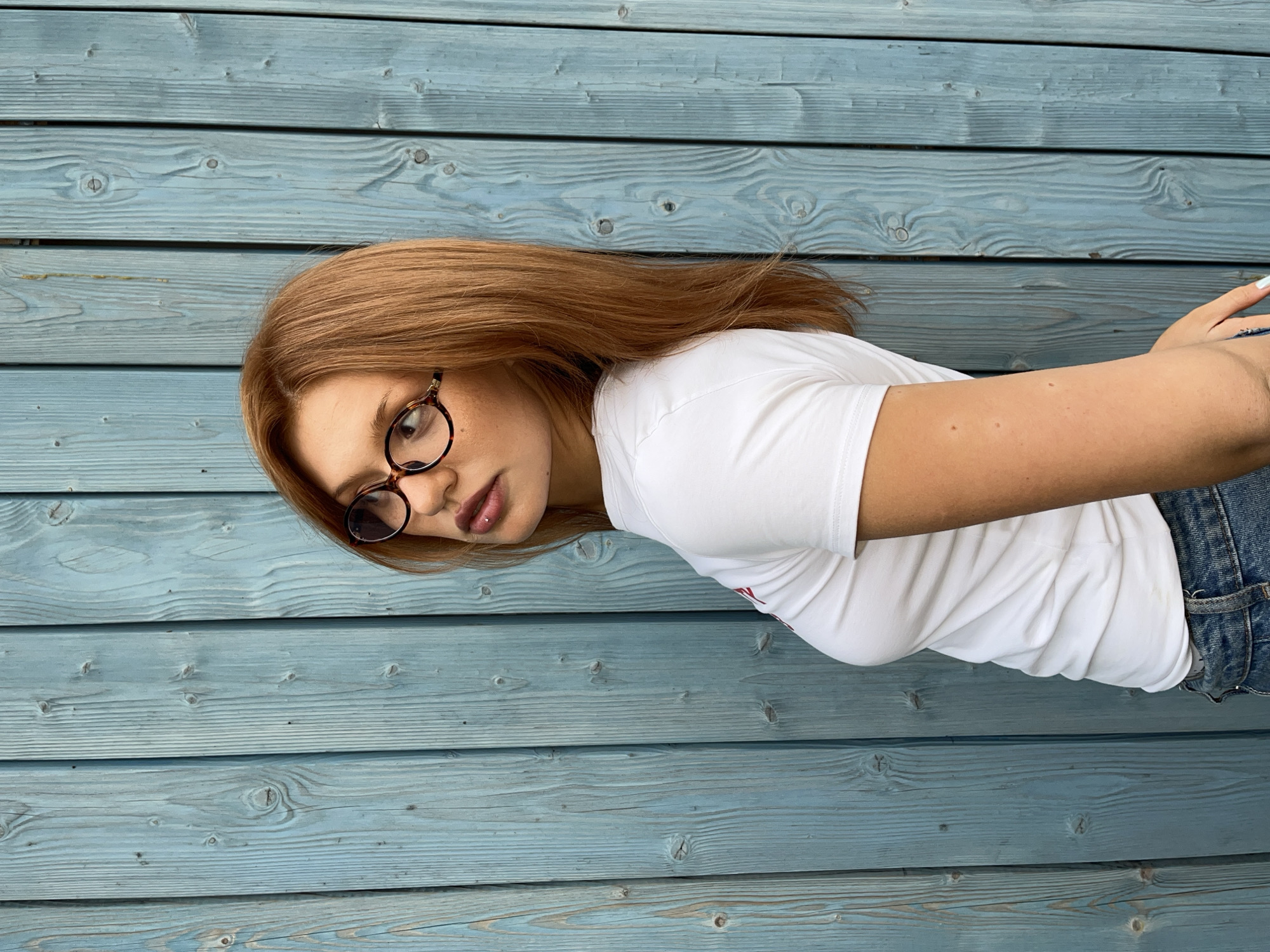 The image depicts a young woman standing against a vertically planked wooden wall, painted in a desaturated blue-green or teal color that reveals prominent wood grain and knots. The woman, appearing to be a young adult with shoulder-length, reddish-brown hair, is positioned with her body angled approximately three-quarters away from the viewer, looking back over her left shoulder. She wears round, tortoiseshell-patterned eyeglasses. A small, metallic stud piercing is visible in her philtrum, above her slightly parted lips. She is dressed in a fitted white short-sleeved t-shirt and blue denim jeans. Her right arm and part of her hand, showing light-colored nail polish, are visible near her hip. The scene is illuminated by soft, diffuse lighting. The specific location within Chaikovsky, Russia, cannot be determined from the visual information provided.