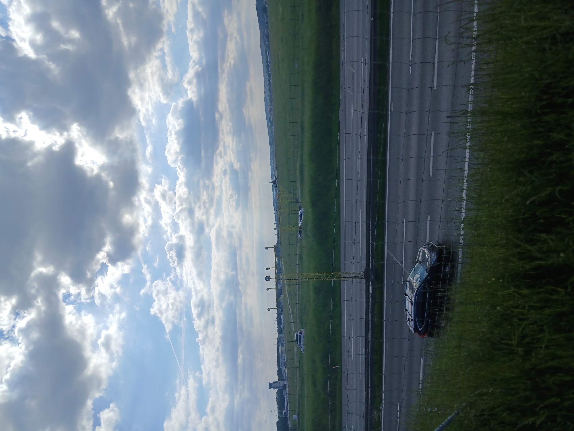 The image shows a view of a highway from an elevated position, likely a bridge or overpass. The sky is filled with cumulus clouds. A section of green grass separates the highway from a chain-link fence. A dark-colored car is parked partially on the side of the road, near the grass. In the distance, there are airport lights and buildings along the highway, suggesting the highway is located near an airport.