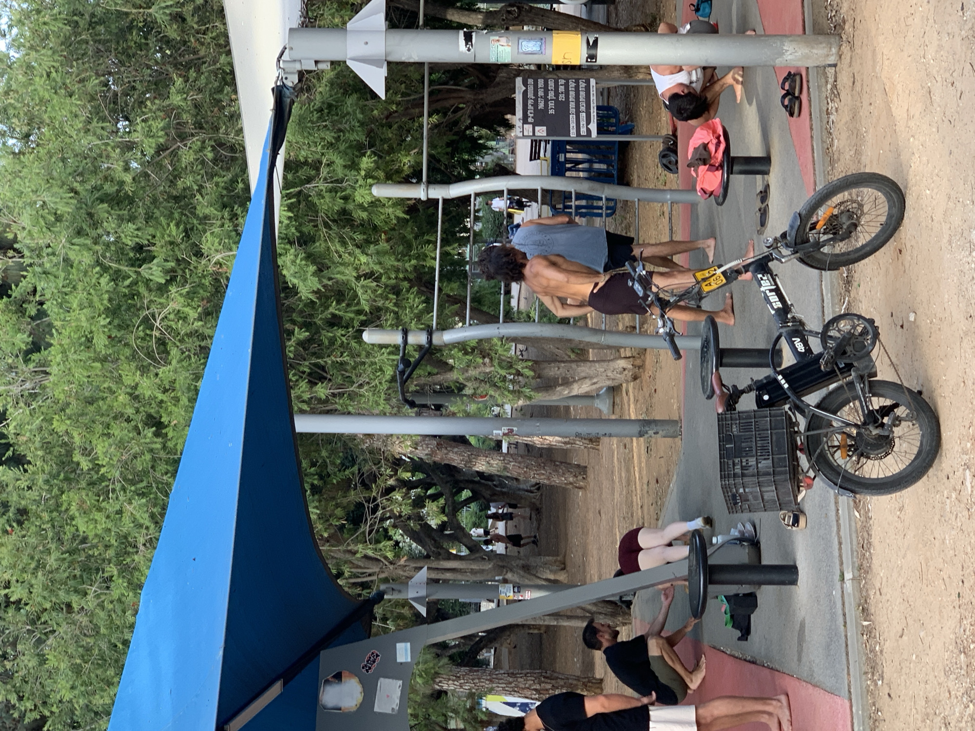 The image shows an outdoor exercise area in Tel Aviv, Israel. Several people are engaged in various workout activities, including using exercise machines designed for core strengthening. An electric bike is parked nearby. A blue sunshade offers partial cover to the area. Trees line the perimeter of the workout space, and signage with Hebrew text is visible on a post. The location details, including street names and regulatory information, are partially legible on the signage, suggesting it is a well-maintained public fitness area in Tel Aviv.