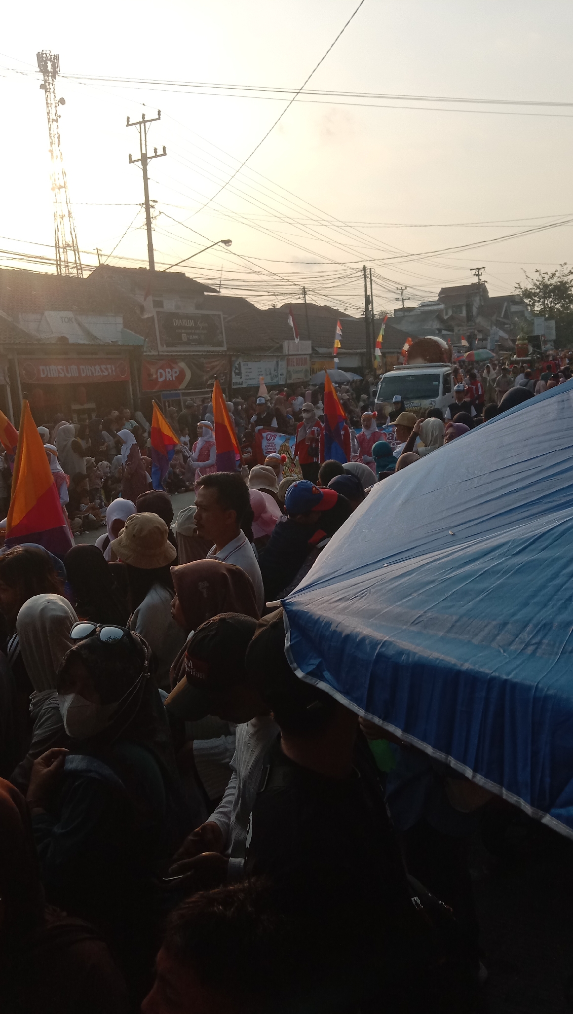 The image depicts a street scene in Jatisawit, Indonesia, during what appears to be a daytime event with a crowd gathered. The lighting suggests either early morning or late afternoon, with the sun low in the sky, casting a warm glow. In the background, a tall telecommunications tower stands prominently to the left, alongside utility poles and a dense network of overhead wires. Several buildings with tiled roofs line the street, some displaying signage, including "DJARUM" and "DIMSUM DINASTI."
The foreground and mid-ground are filled with a diverse crowd of people, many wearing casual clothing. Some individuals are holding flags, predominantly orange and red, which are visible throughout the scene. A large blue tarp, possibly an umbrella or canopy, dominates the right side of the image, partially obscuring some of the people and objects behind it. People's faces are partially visible, and many appear to be looking in the same direction, suggesting they are watching a procession or event. There is a white van or truck visible in the mid-ground. The overall atmosphere seems to be that of a community gathering or a public event.