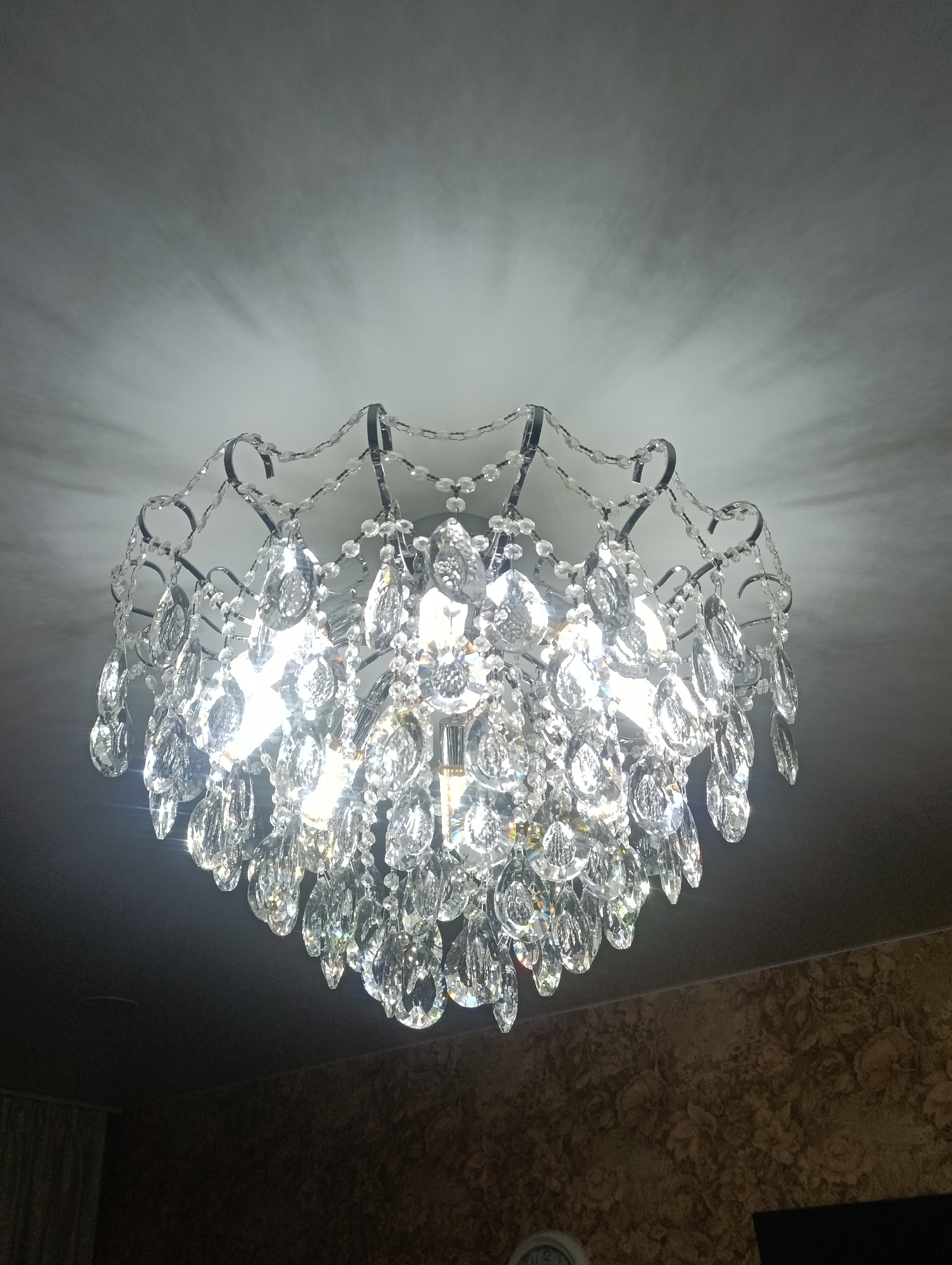 The image shows a chandelier hanging from a ceiling. The chandelier is ornate, with a dark-colored metal frame and numerous clear crystal pendants. The ceiling is white, and the chandelier's light creates a starburst effect on the ceiling. In the background, the lower portion of a patterned wall is visible along with a clock. The scene is likely indoors. There is no evidence to suggest that the location is in Krasnoyarsk, Russia.