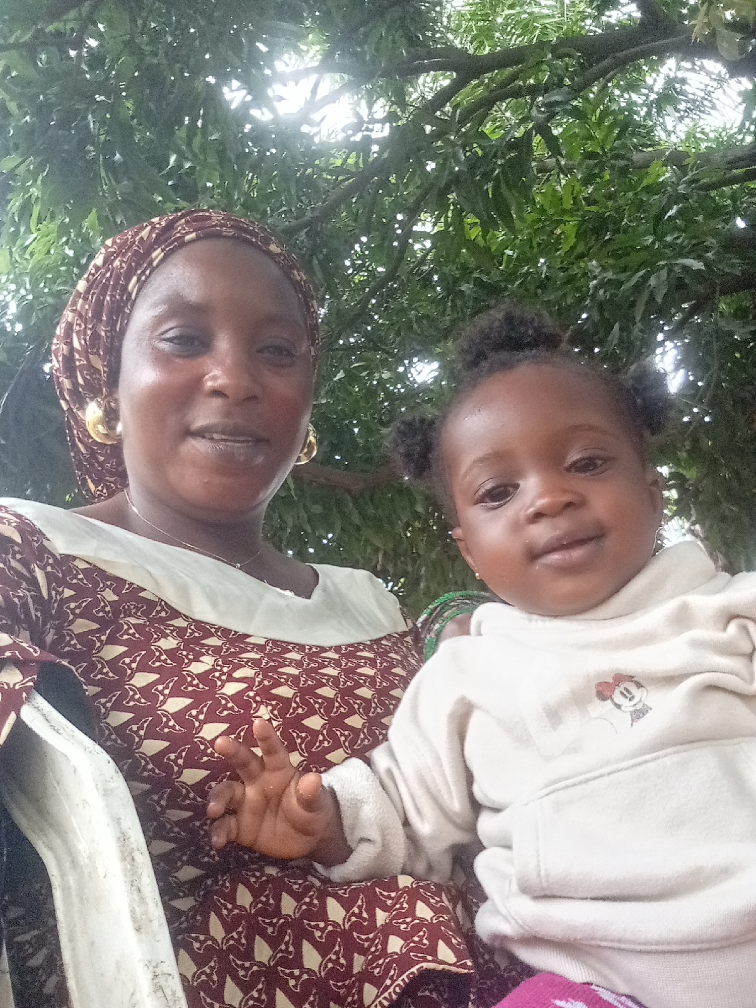 The image features an adult woman and a young child posing for a close-up photo outdoors in Andaha, Nigeria. The woman, with a medium-dark complexion, wears a patterned maroon and off-white head covering and a matching top with a plain light-colored upper section. She has large gold-colored hoop earrings and a thin silver-colored necklace, and her expression is a gentle smile.
The child, likely a toddler, has a dark skin tone and hair styled in two small puffs. They are wearing a cream or off-white hooded sweatshirt with a small, embroidered Minnie Mouse character visible on the chest. The child looks towards the camera with a neutral, slightly curious expression, and a small hand is visible, resting near the woman's patterned attire.
In the background, lush green foliage and tree branches are visible, with bright, diffused light filtering through the leaves, indicating a daytime outdoor setting, possibly under a shaded tree canopy. Part of a white chair or seating surface is visible on the left side of the frame, supporting the woman. The overall impression is one of calm and warmth. No explicit text is visible, only the Minnie Mouse character design.