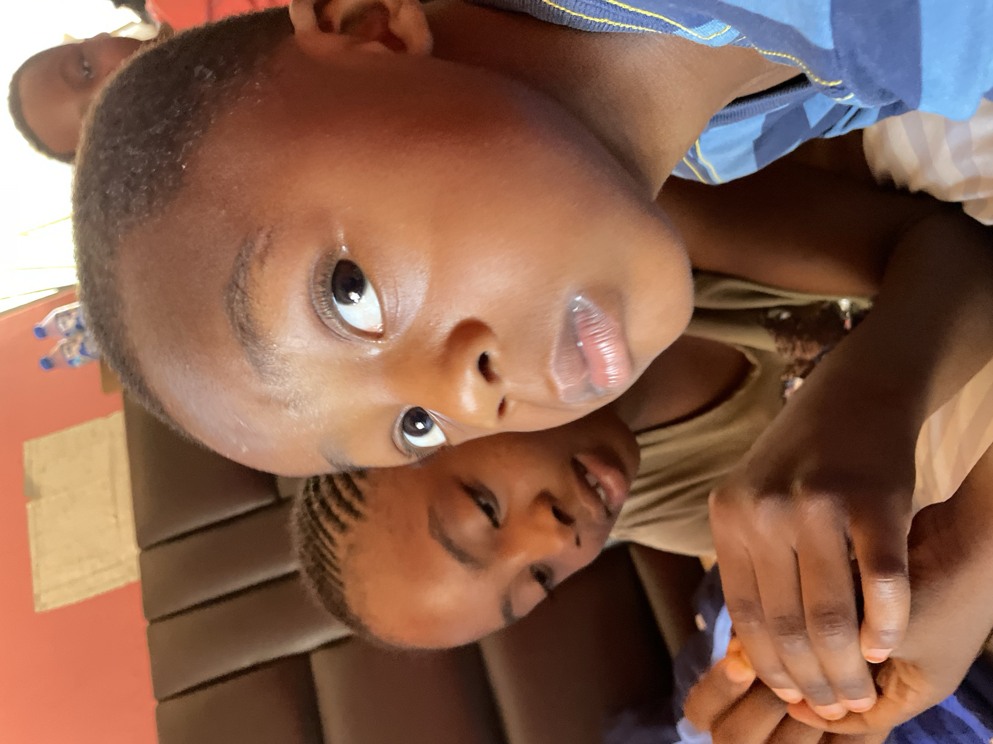 Two young, dark-skinned boys are closely positioned in the frame. The boy in the foreground is wearing a blue striped shirt, his face is the focal point. The other boy is slightly behind, in a brown shirt. A third boy appears faintly in the upper left corner. They are in an interior setting, possibly a residence, suggested by the brown horizontal slats behind them. The specific location is stated as Jos, Nigeria, though this cannot be verified from the image. The lighting seems natural.