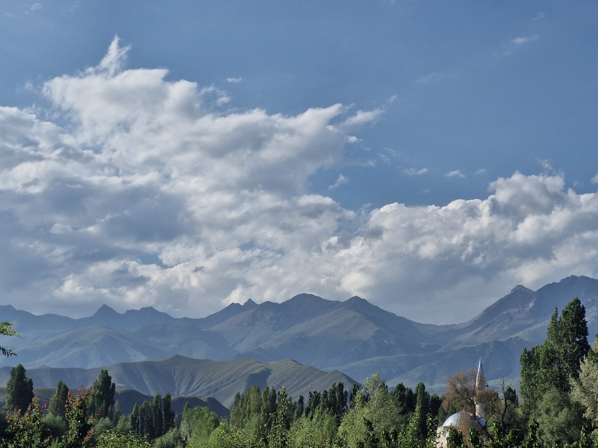 The image depicts a serene landscape with a prominent mountain range in the background under a partly cloudy sky. In the foreground, a lush forest of green trees occupies the lower portion of the frame. Partially visible within the trees, on the right side of the image, is a mosque with a dome and a minaret, suggesting a location in a rural or semi-rural area. The mountainous terrain is characterized by layered peaks that recede into the distance, with varying shades of blue and green indicating depth and atmospheric perspective. The sky is a vibrant blue, interspersed with fluffy white and gray clouds that cast shadows on the mountains. The overall scene conveys a sense of tranquility and natural beauty. The image is assumed to be of the city of Bulan-Segettu, Kyrgyzstan, based on the provided context.