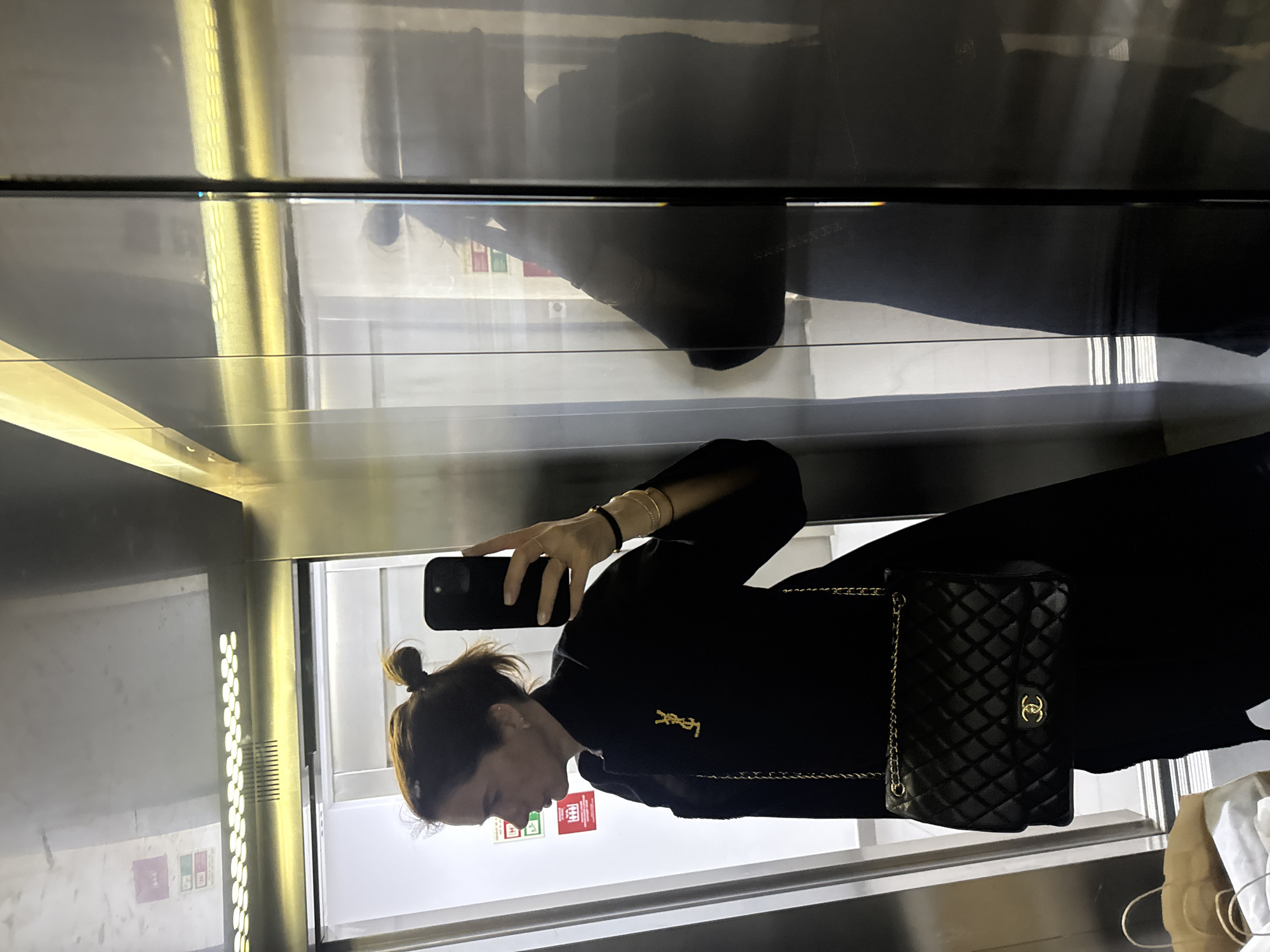 A woman is taking a selfie in an elevator with mirrored walls. She is wearing a black outfit, including what appears to be a black dress or top, and a black quilted Chanel bag. She has her hair in a bun and is holding a smartphone in her right hand to take the picture. She has multiple bracelets on her left wrist and a YSL logo is prominently displayed on her outfit.
The elevator interior is metallic, with vertical light panels. The reflection in the elevator shows the surroundings, including a wall with what appear to be safety information posters or notices. Based on the signage visible on the elevator wall and the overall architecture, the location is likely within Lisbon, Portugal.