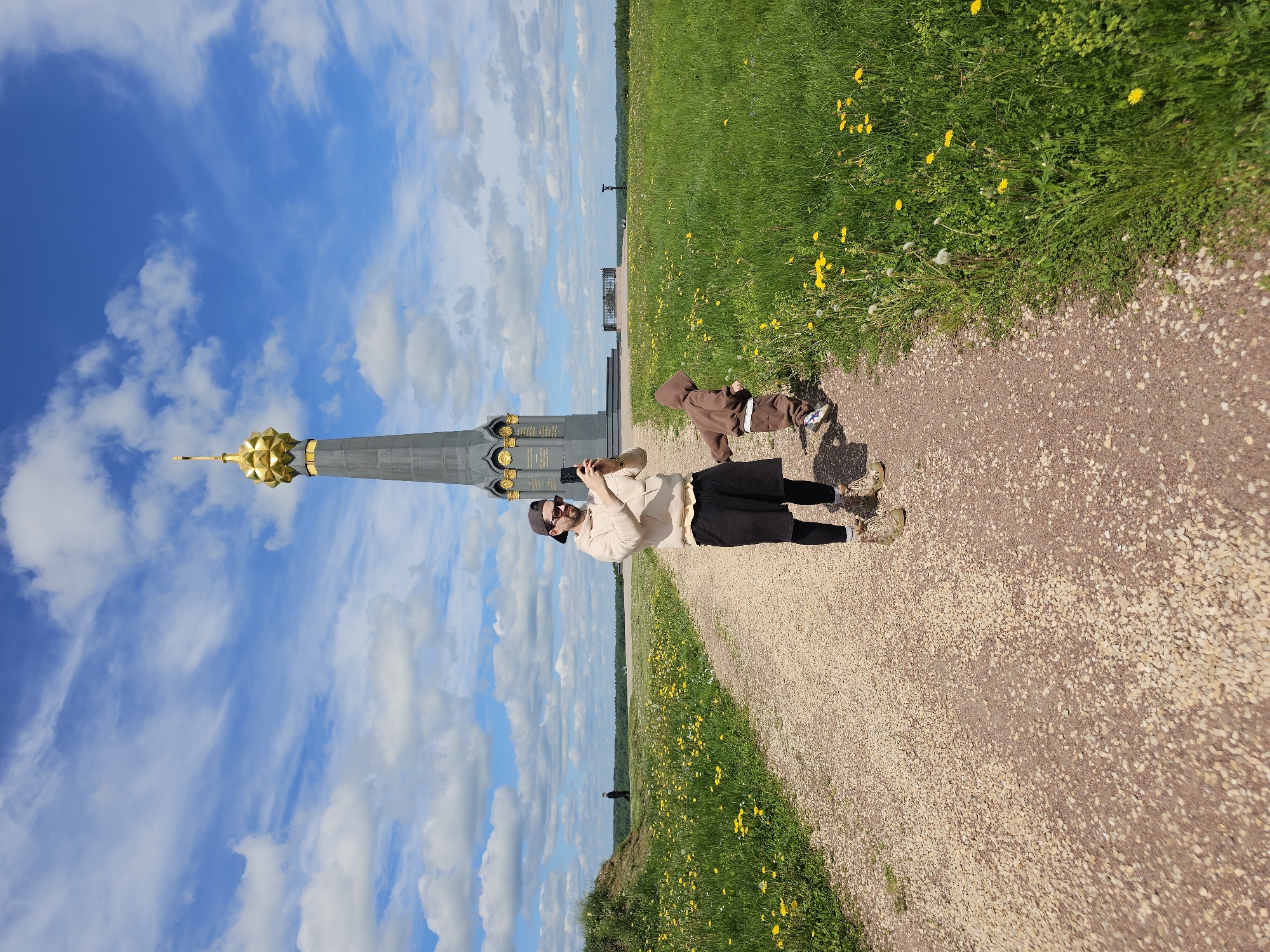 The image shows a man and a child standing on a gravel path in an outdoor setting. The man is wearing a beige jacket, black pants, a cap, sunglasses, and hiking boots. He is holding a smartphone, presumably taking a photo of the large monument located beside him, which features a golden dome on top. The child, wearing a brown hooded outfit, is standing to the right of the man. The scene takes place on a path flanked by green grassy areas dotted with yellow wildflowers under a blue sky filled with white clouds. The location is in the city of Unknown, Unknown.