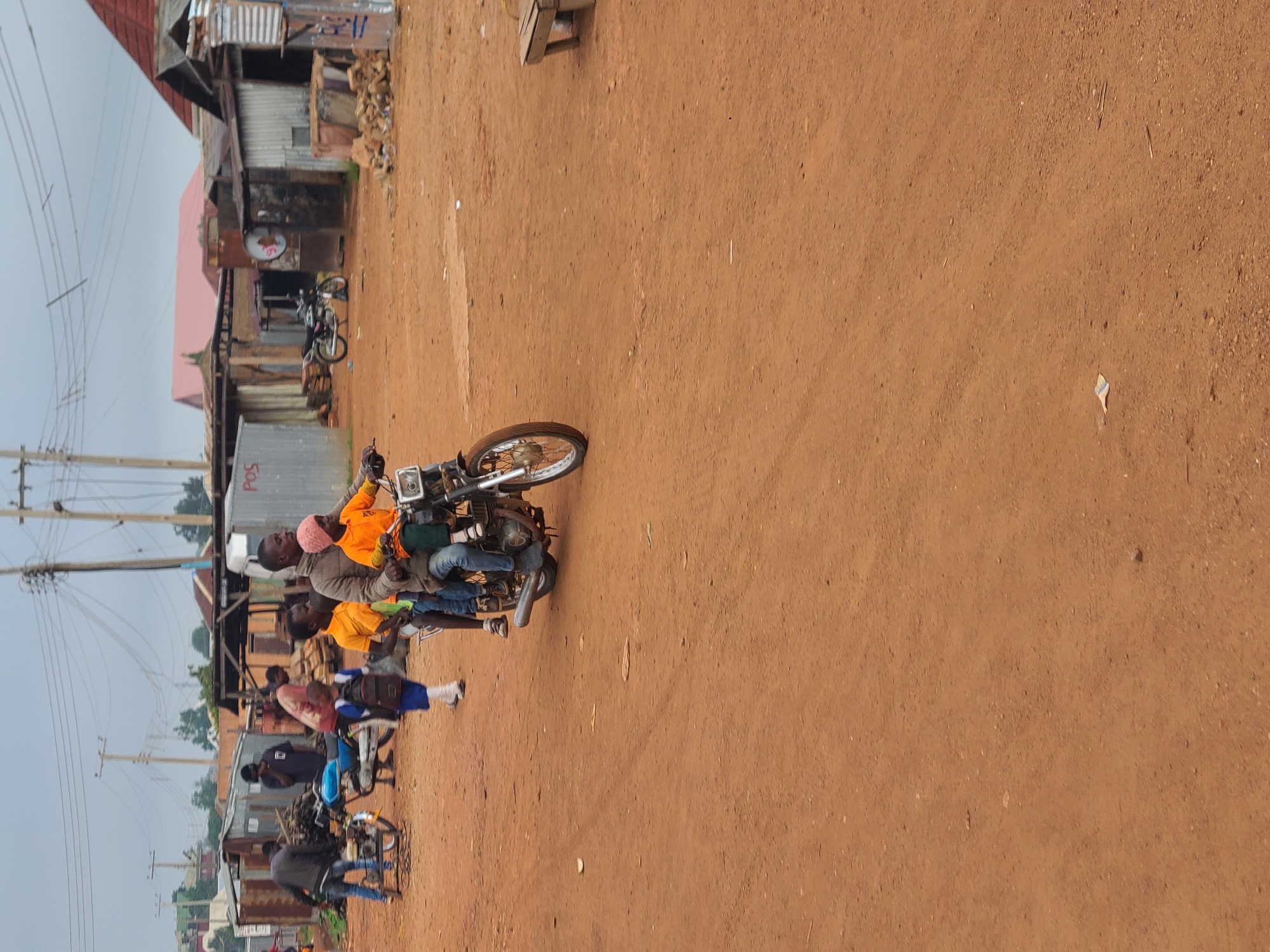 A group of people are riding on motorcycles in a rural area in Barikin Sabon Gidan Kanat, Nigeria. The scene depicts a dusty, unpaved road with simple, rustic buildings lining the side. Several motorcycles are visible, carrying multiple passengers. In the foreground, one motorcycle has three people on it, including a driver and two passengers. They are dressed in casual clothing. Further in the background, other motorcycles are also carrying passengers, suggesting a form of local transportation or a gathering. The buildings appear to be made of corrugated metal and wood, with some appearing to be shops or residences. There are power lines and utility poles visible, indicating the presence of electricity. The ground is a reddish-brown dirt, common in many parts of Nigeria. The sky is overcast, suggesting it might be a cloudy day. The overall impression is a snapshot of daily life and transportation in a rural Nigerian community. A sign with "POS" is visible on one of the buildings, likely indicating a point of sale or service. The image seems to be taken during daylight hours.