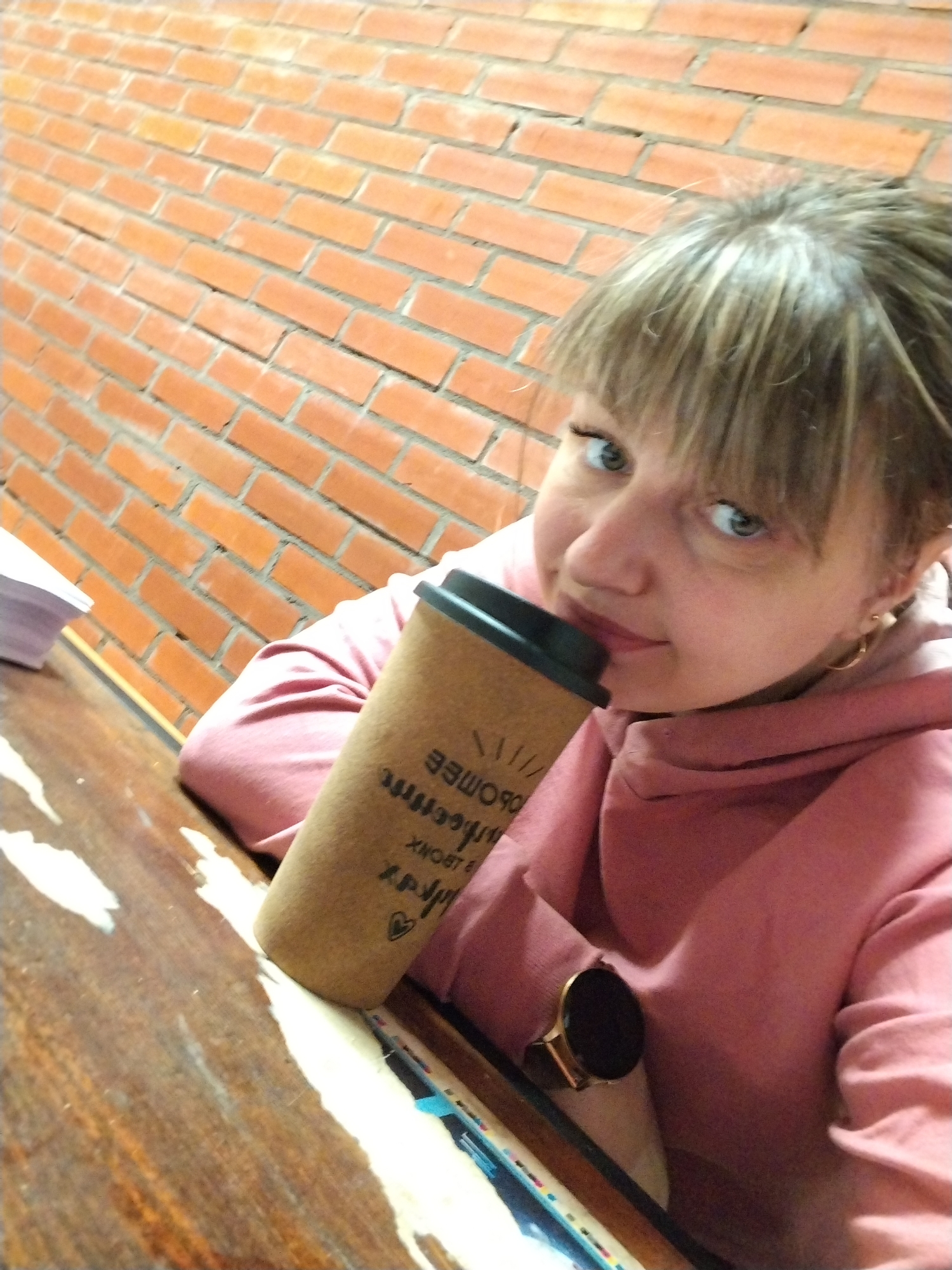 The image captures a woman indoors, likely in a casual setting such as a café or public space in Шебунино, Russia. The primary subject is the woman, who appears to be taking a selfie from a slightly high angle, tilting her head. She has light brown hair with bangs, light blue or green eyes, and a friendly, engaging expression as she looks directly at the camera with a subtle smile. She is wearing a pink hooded sweatshirt and small hoop earrings, and a dark smartwatch with a gold-colored frame is visible on her left wrist.
Her arms are resting on a prominent brown wooden table with visible wear, featuring lighter streaks and patches. On the table to her left, a stack of white papers is visible. She is holding a light brown, cylindrical disposable coffee cup close to her face with both hands.
The background features a distinct red-orange brick wall, providing a warm, textured backdrop. The lighting suggests an indoor environment.
The coffee cup has reversed black text and graphic elements printed on its side. When mirrored, discernible Russian words include "ПРОМЫСЕЛЪ" (an archaic form of "craft" or "trade") and "КОФЕ" (coffee), along with other less clear text, a small heart icon, and a design resembling radiating lines. The overall ambiance of the image is casual and relaxed.