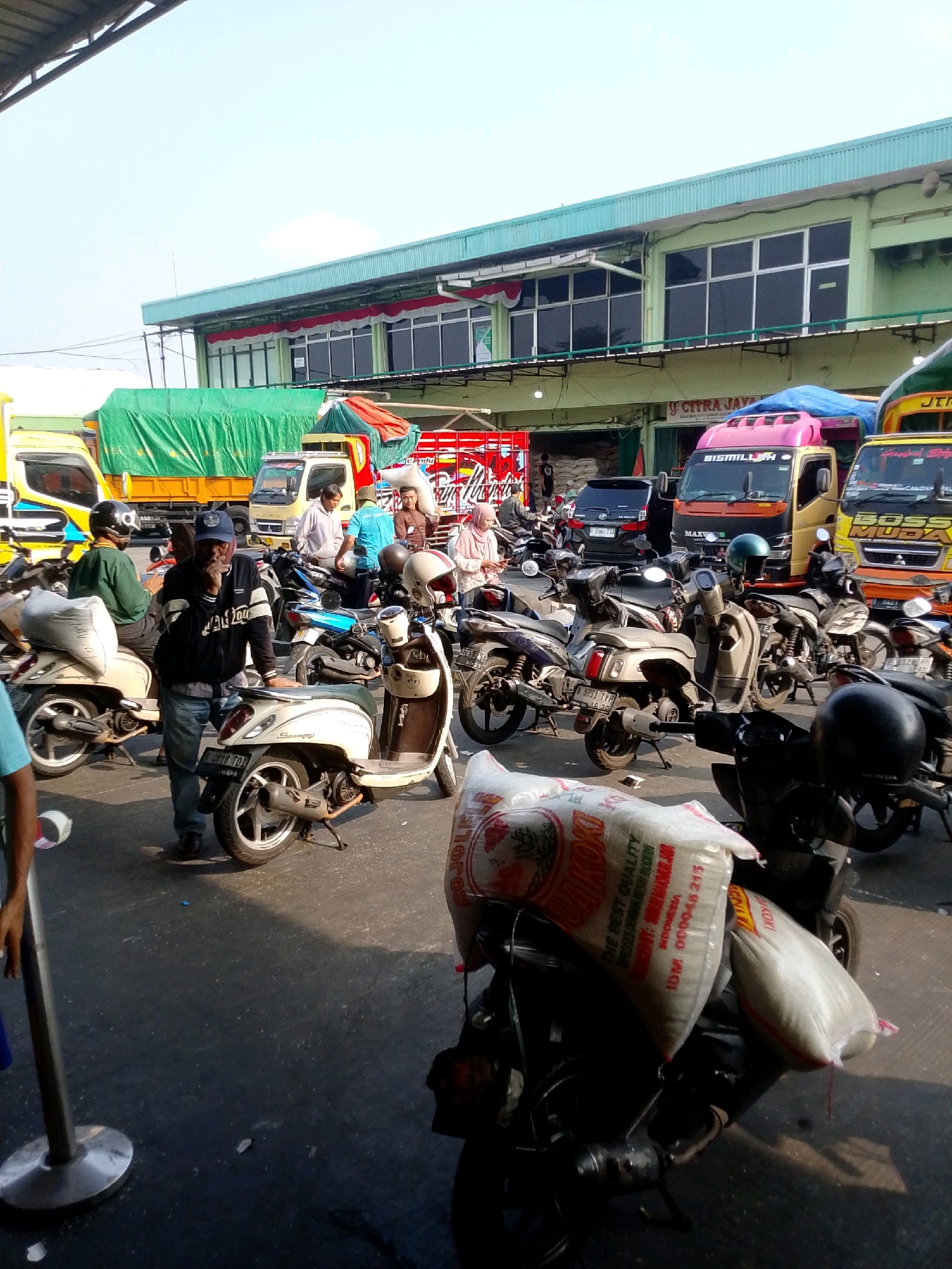 This is a horizontal, medium-shot, eye-level photograph taken outdoors on a sunny day. The scene depicts a bustling market or loading area, likely in the city of East Jakarta, Indonesia, given the presence of Indonesian text on the trucks and bags. In the foreground, a man in a blue baseball cap and black jacket is standing next to a cream-colored scooter, smoking a cigarette and looking downwards. Several other motorcycles and scooters are parked around him, some carrying large sacks of what appears to be rice. In the midground, there are multiple trucks with colorful branding and tarpaulins, suggesting they are used for transport and possibly agricultural goods. A few other people, including men and women, are visible in the background, some walking and others near the trucks. The environment appears to be an open-air space with a building in the background that has green and teal colored awnings and large windows. The ground is asphalt. The overall impression is one of a busy commercial hub.