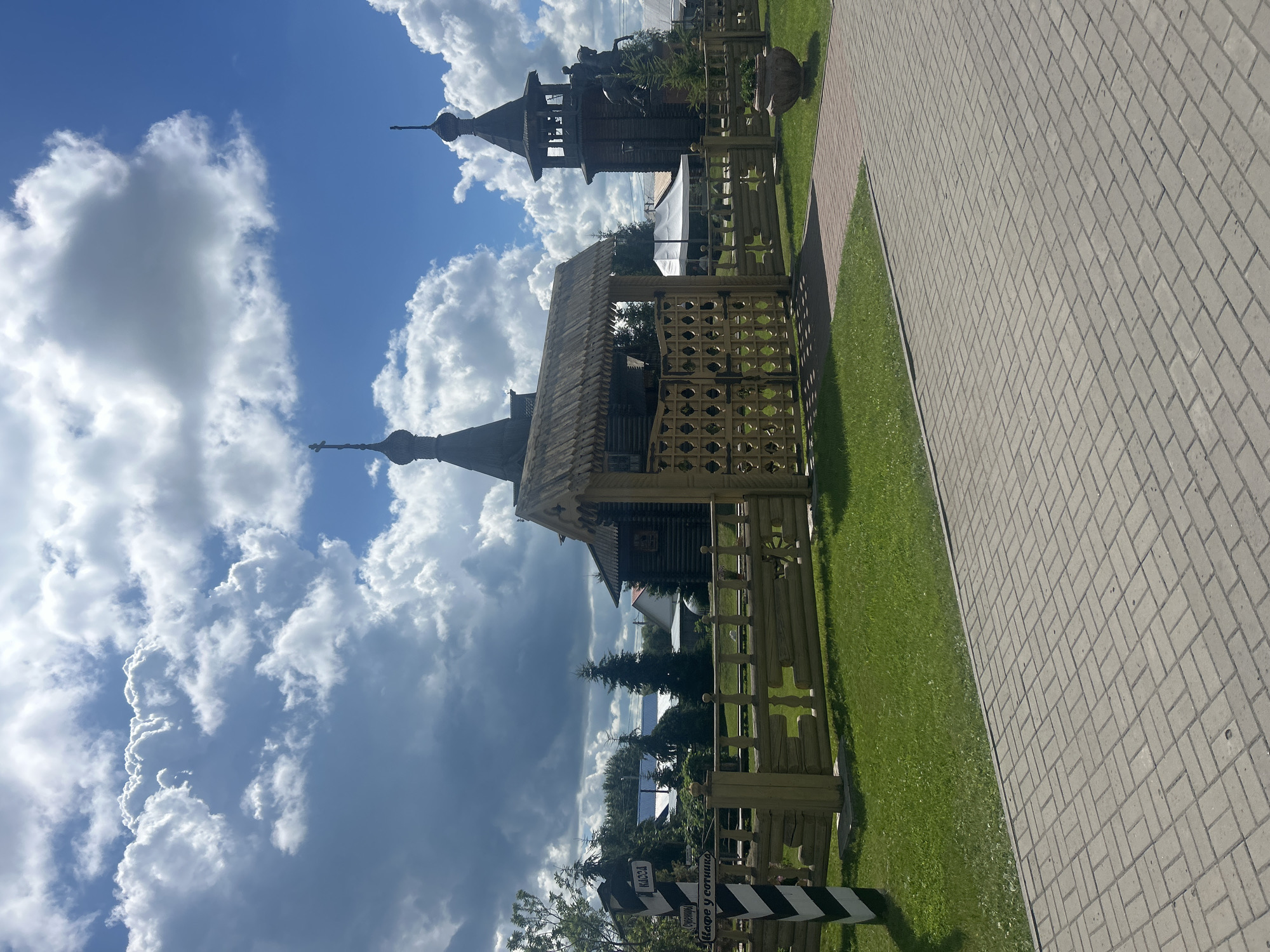 In Bolsherechye, Russia, a traditional wooden gate with a thatched roof stands open, leading to a fenced area. Beyond the gate, a wooden structure resembling a church with a dark, conical roof and a cross is visible. To its right, a wooden bell tower with a similar roofline stands tall, featuring a statue of a man on horseback at its base. A paved walkway leads towards the gate, bordered by a neatly manicured lawn. A signpost with black and white stripes indicates directions to "KACCA" (cashier), "Сувениры" (souvenirs), and "Кафе у сотника" (at the centurion's cafe). The sky above is a vibrant blue, dotted with large, fluffy white clouds. The scene is peaceful and evokes a sense of historical or cultural significance within the town.