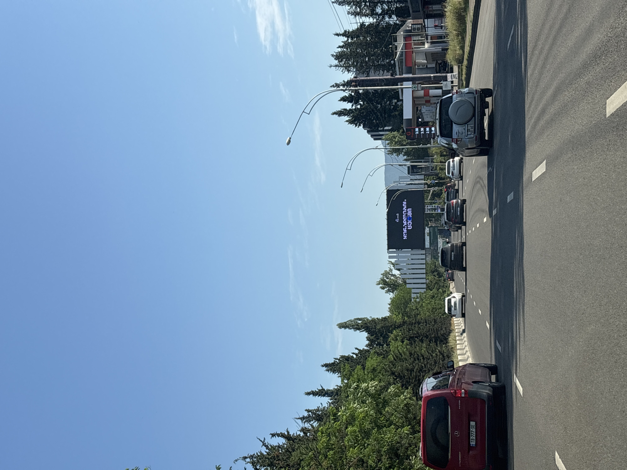 This is a horizontal, eye-level, medium shot of a road in Tbilisi, Georgia on a clear, sunny day. The road appears to be a multi-lane highway or major city street with a clear blue sky above. Several cars are driving on the road. On the left side of the road, there is a dense line of tall green trees. On the right side of the road, there is a gas station with a white canopy and red accents, and several more trees. A large electronic billboard with text in a foreign language is visible in the distance. The overall scene depicts a typical urban driving environment.