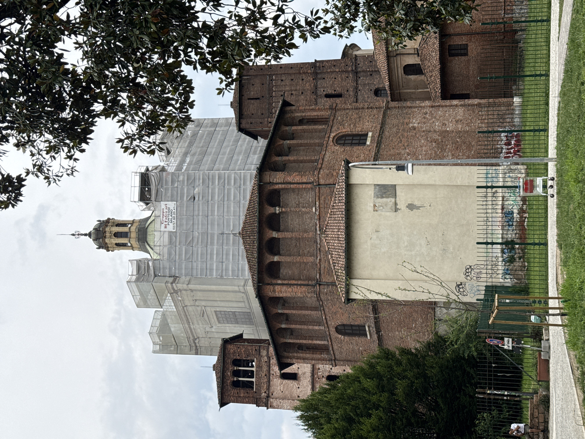 The image captures a historical brick building, likely a church or basilica, in Milan, Italy, largely undergoing renovation. The structure, featuring traditional brickwork, arched windows, and corbels, is partially covered by extensive white scaffolding and netting. A distinctive cupola with a spire crowns the building, also partially obscured by scaffolding.
In the foreground, lush, dark green foliage from broad-leaved trees frames the upper left of the scene, while a dense green bush occupies the lower right. To the right of the main building, a paved pathway runs alongside a grassy area, separated by a dark metal fence. Behind this fence, a light-colored wall is heavily adorned with various graffiti tags and drawings.
Notable details include an overcast sky, suggesting a cloudy day. A silver lamppost stands near the fence, and a discarded shopping cart is visible close by. Street signs, including a circular red one with a diagonal line (likely "No Entry" or "No Stopping"), are also present. A white sign on the scaffolding displays "Gruppo S.r.l. di Falco MILANO" along with the phone number "02/57 60 25 41 A," identifying the renovation company. A lone individual is faintly visible sitting behind the fence in the distance.