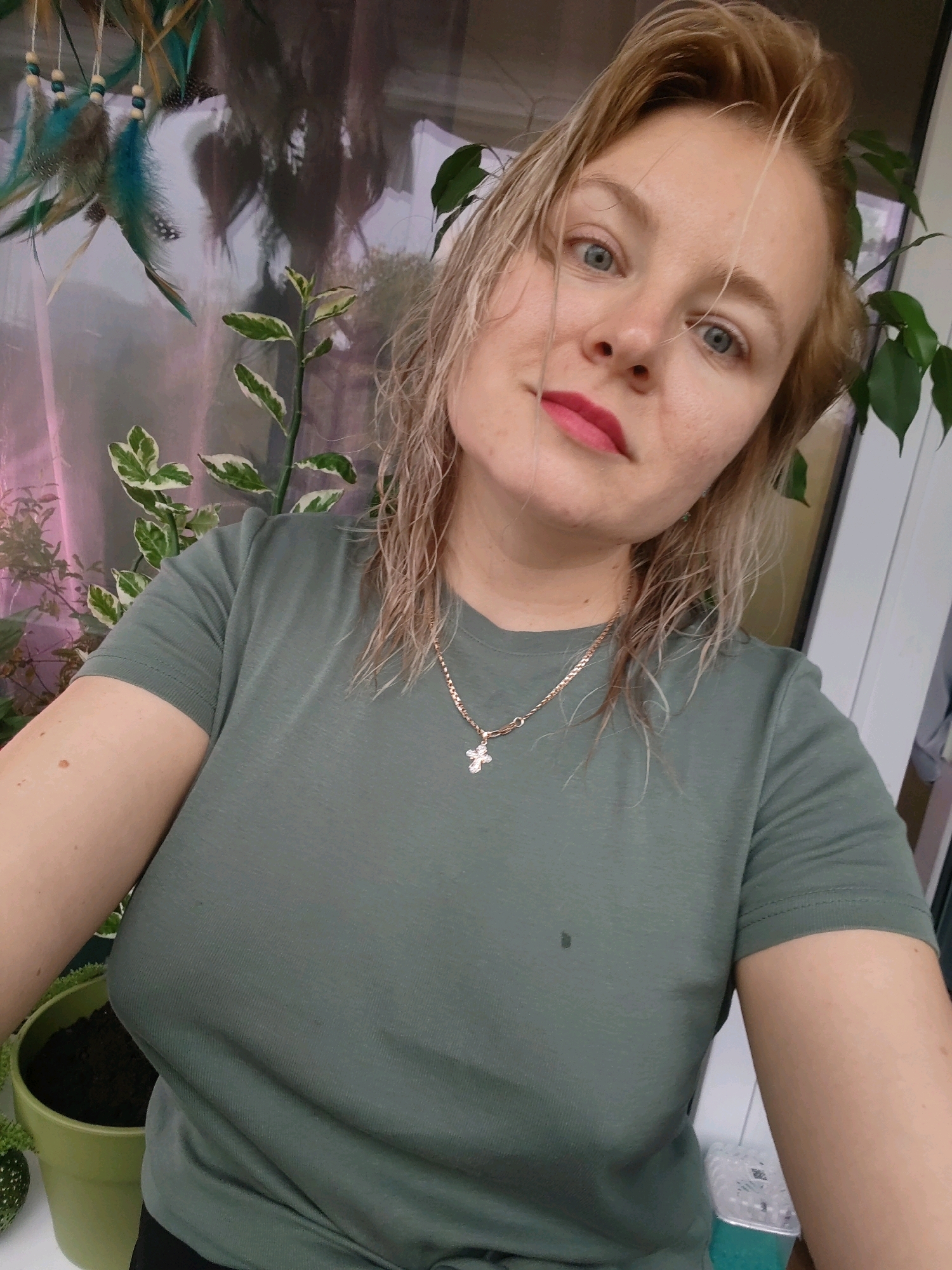 A woman with blonde, wet hair, wearing a green t-shirt and a cross necklace, poses for a selfie indoors. She has blue eyes and pink lipstick. In the background, a plant with variegated leaves and a dreamcatcher with feathers are visible. A green pot with soil is on the left side of the image. The scene appears to be an apartment or house, possibly in Izhevsk, Russia.