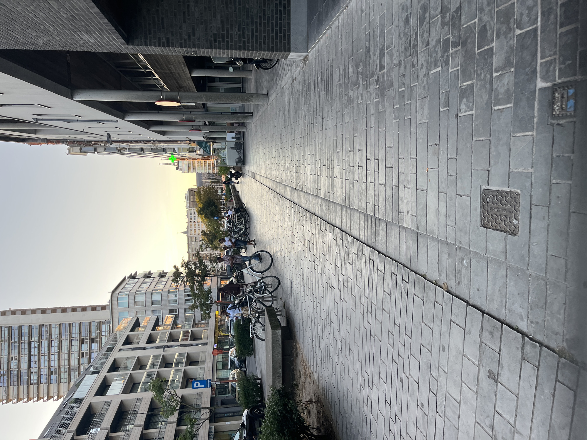The image depicts a street scene in Ostend, Belgium, during what appears to be late afternoon or early evening. A paved walkway, laid with large, rectangular grey stones, dominates the foreground and extends into the distance. The walkway is divided by a slightly raised seam. To the left of the walkway is a patch of greenery and a concrete bench, with several bicycles parked nearby. Beyond this, cars and buildings are visible.
Several people are present on the left side of the street, near the parked bicycles. One individual appears to be talking on their phone while standing next to a bicycle. Others are interacting near a row of shops and cafes lining the street. The buildings on the left are modern, multi-story structures with balconies and large windows, suggesting a residential or commercial area.
On the right side of the street, a modern building with a dark brick facade and large concrete columns is prominent. A motorcycle is parked in front of this building. Further down the street, more buildings and trees can be seen. The sky is clear and shows a warm, golden hue near the horizon, indicative of sunset. The overall impression is a lively urban environment with a mix of modern architecture and pedestrian activity.