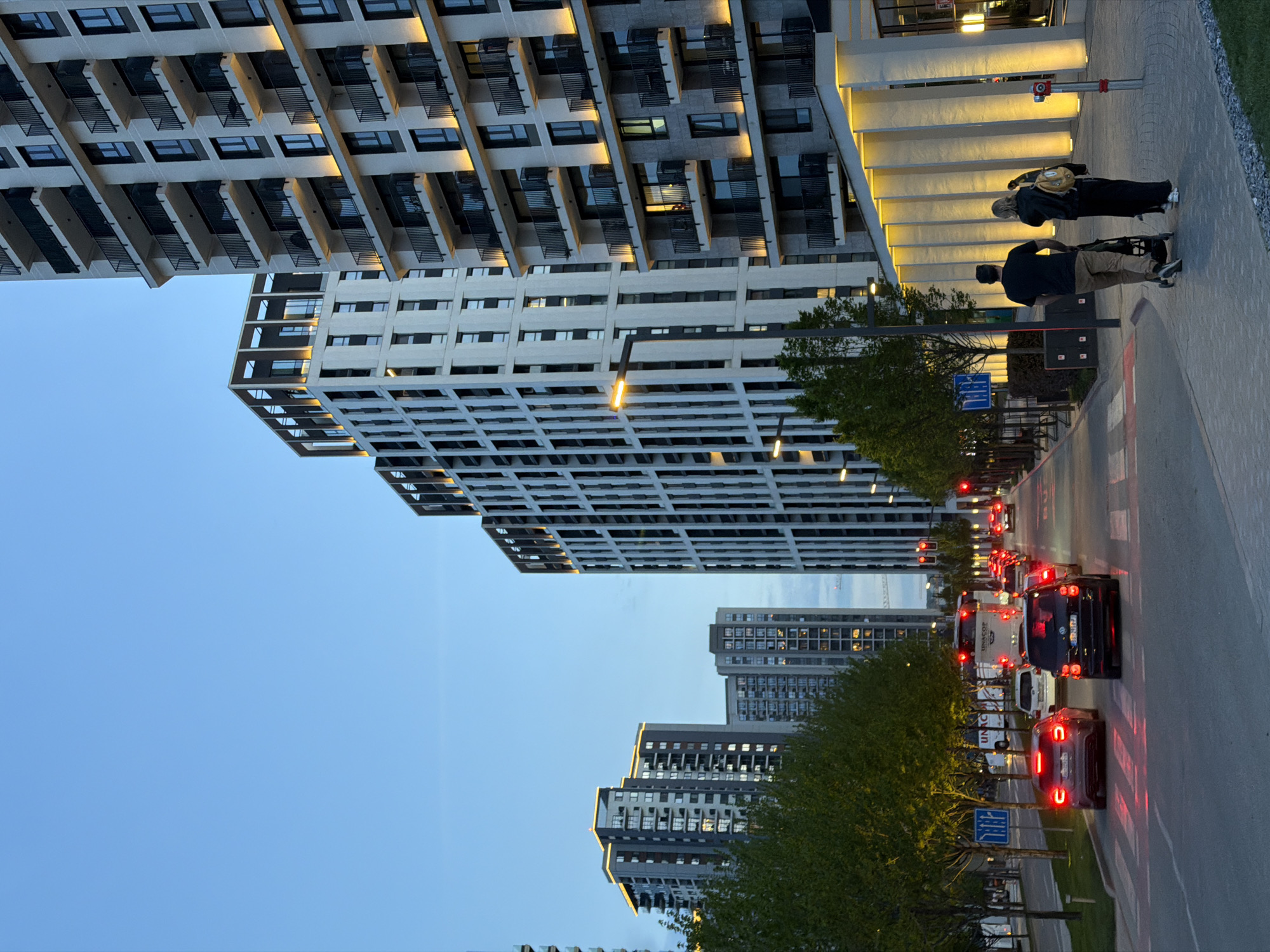 The image depicts a street scene in Belgrade, Serbia. High-rise buildings, of modern architectural design, dominate the view. A pair of pedestrians are walking along a sidewalk adjacent to the street. A line of cars with lit brake lights indicate halted traffic. Trees line the street separating the sidewalk from the vehicle lanes. The sky is a gradient of light blue.