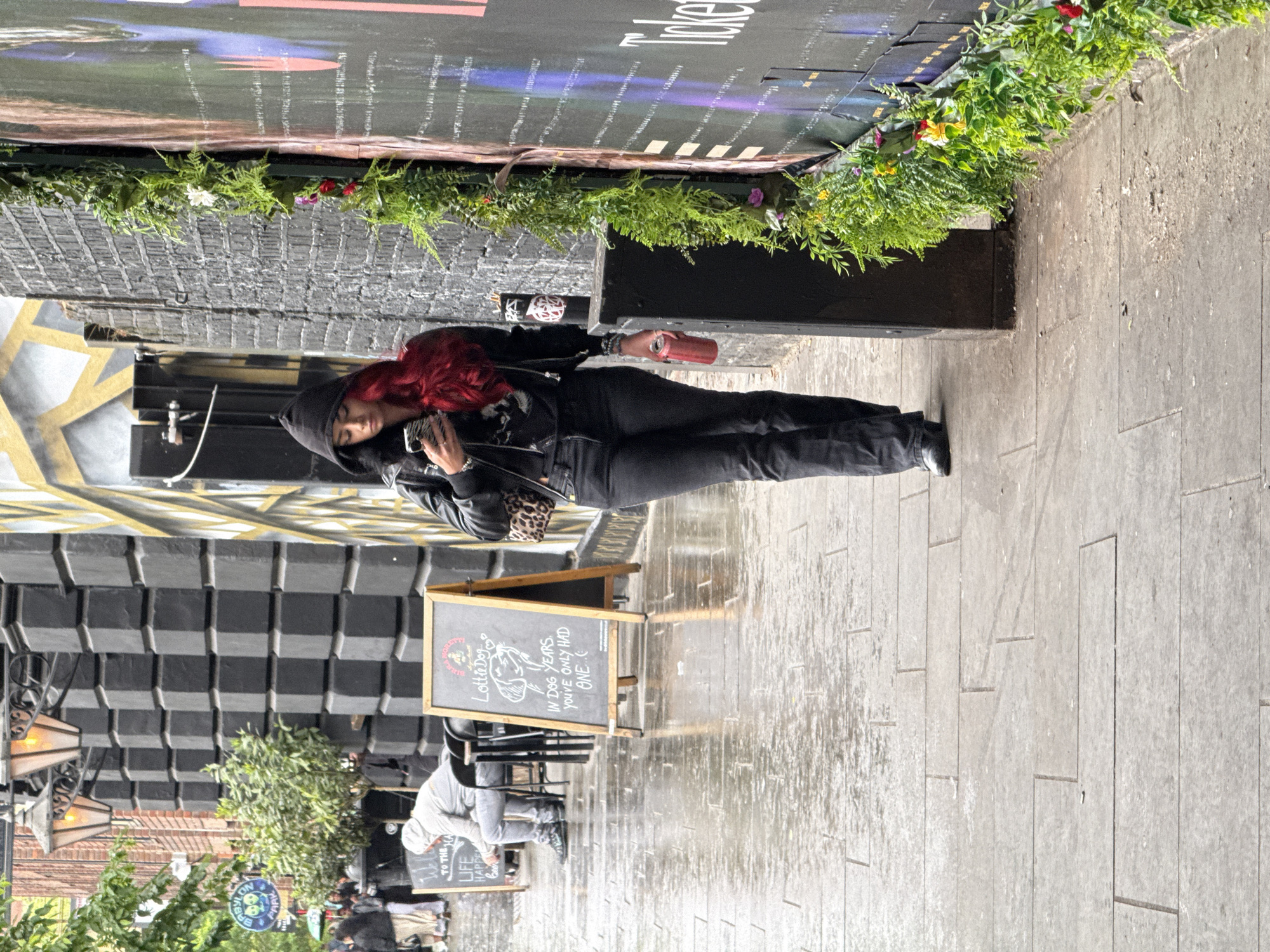 A street scene is captured with a woman as the primary subject, standing on a sidewalk in what appears to be the city of London, United Kingdom. She is wearing a black hooded jacket and dark clothing, holding a phone and what seems to be a canned beverage. She has bright red hair that stands out against her dark attire.
The sidewalk is paved with gray rectangular stones, and there are signs of moisture, suggesting it may have recently rained or is currently raining. In the background, there's a building with a distinctive facade featuring black and yellow blocks or geometric patterns. A sign in front of the establishment reads "Lottie Dog" followed by the text "In Dog Years you've only had one."
To the left of the woman, there are other people present in the background, but they are not in focus. Street lamps and greenery add to the urban setting. Above her, there's a wall with plants and a poster.