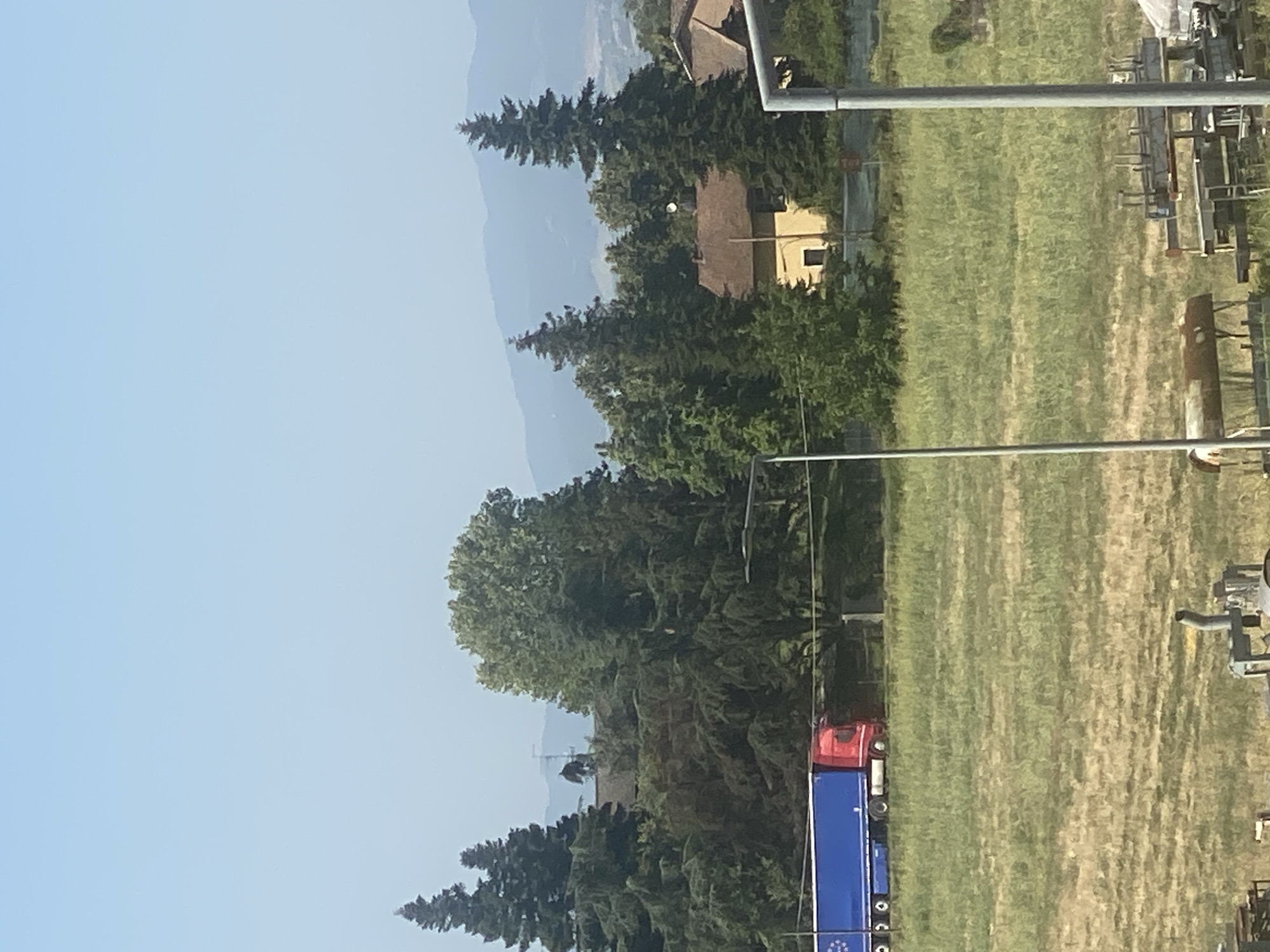 This is an outdoor, daytime, medium-shot photograph taken from a slightly elevated position. The scene depicts a rural or semi-rural area with a clear, pale blue sky. In the middle ground, a bright blue trailer with a red truck cab is driving on a road lined with trees. The truck and trailer appear to be a commercial vehicle. Behind the truck, several evergreen trees and deciduous trees form a dense line. A small, light-yellow building with a brown roof is visible amongst the trees. Further in the background, hazy, blue-tinted mountains are visible on the horizon. In the foreground, a dry, grassy field occupies most of the lower half of the image. Several metal structures, possibly scaffolding or parts of construction equipment, are present in the bottom right and bottom left corners of the frame. There are no discernible people in the image, and no specific landmarks that would definitively place this scene in Parma, Italy. The overall impression is one of a calm, sunny day in a landscape transitioning between agricultural land and wooded areas.