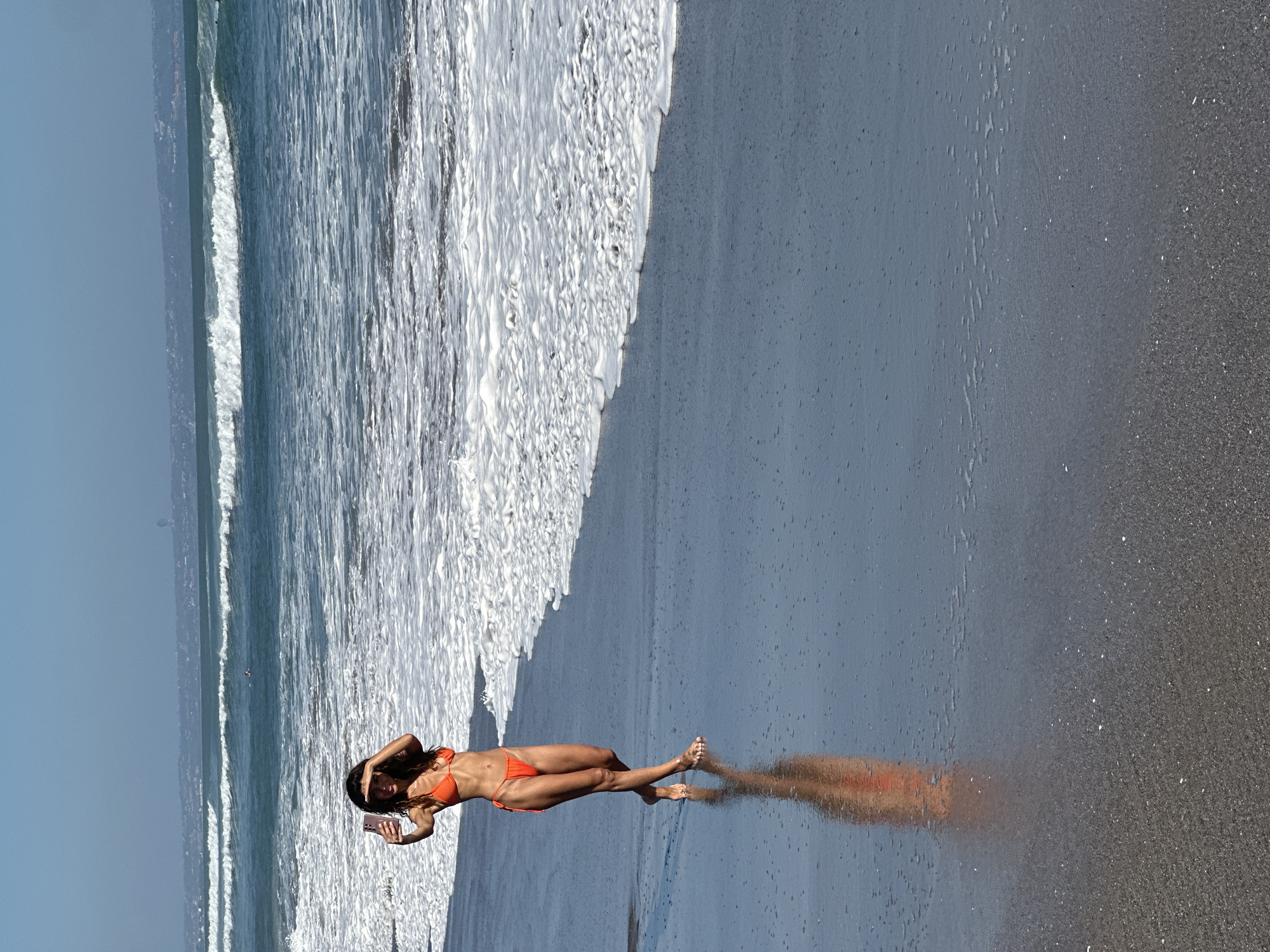 A young woman with long dark hair, dressed in an orange two-piece bikini, walks barefoot on the wet, dark grey/black sand beach in North Kuta, Indonesia. She is positioned towards the left side of the frame, facing slightly towards the viewer. Her left hand holds a pink-cased smartphone at arm's length, angled for a selfie, while her right hand is raised to shield her eyes from the bright sunlight. Her reflection is sharply visible on the highly reflective wet sand. To her right, multiple lines of ocean waves with white foam break towards the shore. In the mid-distance within the ocean, a small dark figure, likely a surfer or swimmer, is visible. A faint, hazy coastline is discernible on the distant horizon under a clear, light blue sky. The foreground consists of textured, wet black sand, displaying small flecks of white material.
