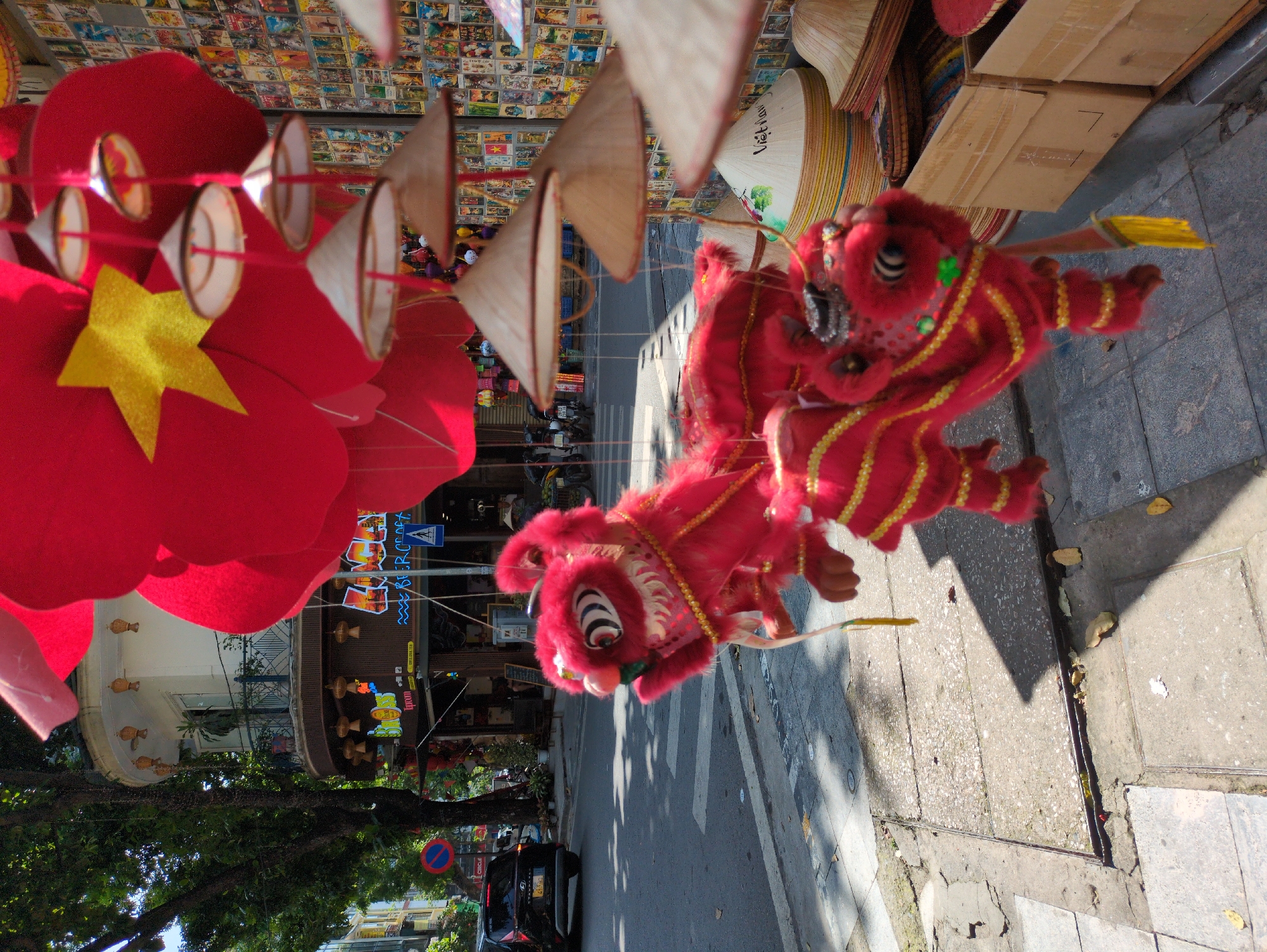 This image captures a street scene in Hanoi, Vietnam, likely during a festive period, given the presence of traditional decorations. In the foreground, two vibrant red lion dance costumes are displayed, adorned with gold trim and tassels. These costumes are suspended from above, suggesting they are part of a market or street stall selling celebratory items. Behind the lion costumes, a series of conical hats, known as "nón lá," are also displayed, some stacked and others hanging, with one prominently featuring the word "Vietnam." The background reveals a bustling street with buildings, trees, and a road where a black car is visible. The architecture of the buildings suggests a mix of older and newer structures. The overall impression is a lively street scene with a strong cultural presence, characteristic of urban areas in Vietnam.