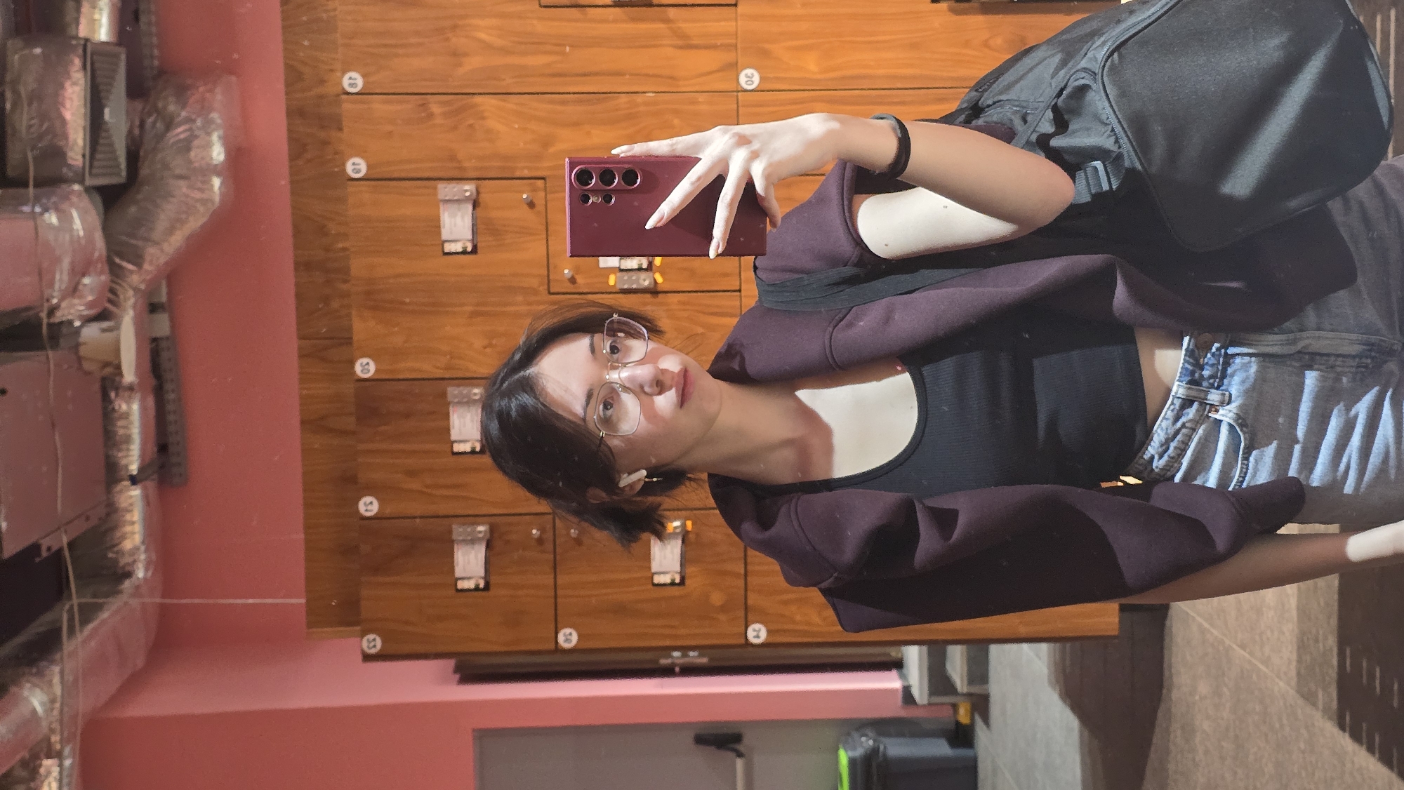 A young woman is taking a selfie in what appears to be a locker room or similar space. She is wearing a black top, denim shorts, and a maroon-colored hoodie. She has glasses, dark hair, and is holding a maroon smartphone in her right hand to take the photo. She has a black backpack slung over one shoulder.
Behind her are rows of wooden lockers with numbered labels. To the left of the frame, there's a pink wall with some ductwork or ventilation system visible. The floor appears to be tiled in light-colored stone. The location cannot be determined to be in Stara Zagora, Bulgaria, from the image alone.