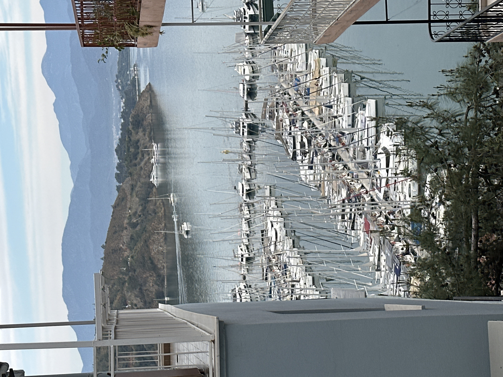 The image shows a view of a marina filled with sailboats, likely located in Fethiye, Turkey, given the presence of a mountainous landscape typical of the region. The foreground includes structural elements of a building, possibly a balcony or rooftop, framing the scene. Numerous white sailboats are docked in the marina, their masts creating a dense network of vertical lines. In the background, there is a body of water with land formations, including a small, tree-covered island. Further in the distance, mountains stretch across the horizon. The sky is lightly colored with a mix of white and pale blue, suggesting either early morning or late afternoon. The overall impression is one of a tranquil harbor scene, blending natural landscapes with human-made structures.