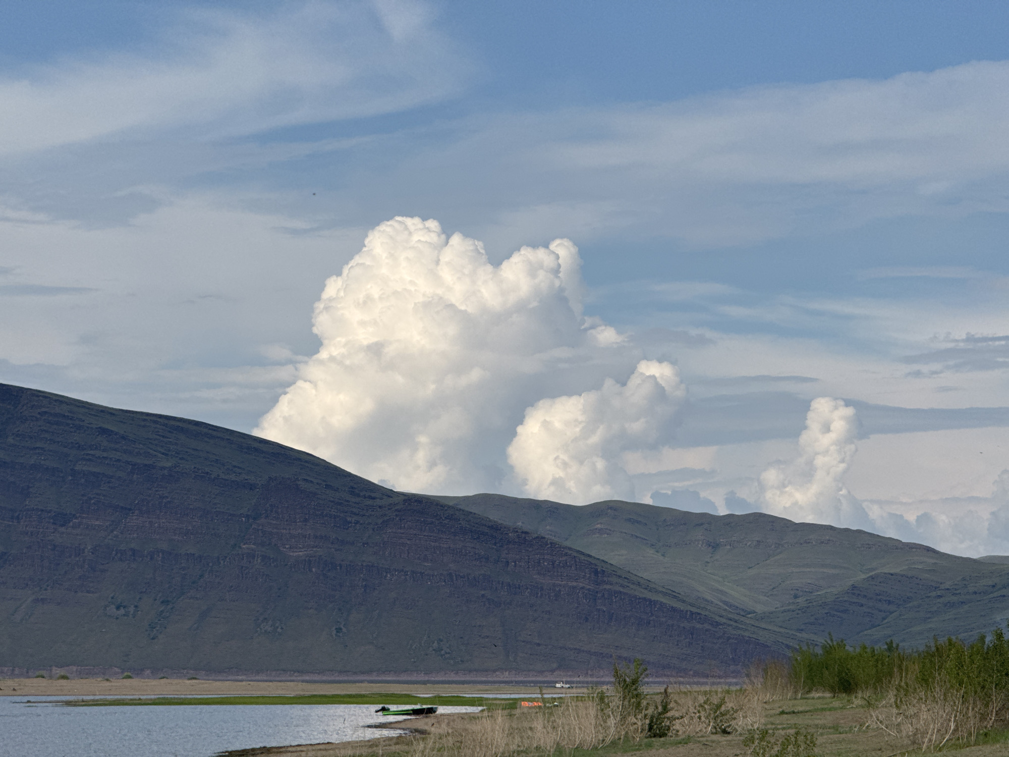A wide landscape photograph captures a body of water in the foreground, bordered by a grassy shoreline. Beyond the shore, rolling green hills rise, with a darker, more rugged mountain range dominating the left side of the frame. The sky above is a blend of blue and white, featuring dramatic, billowing cumulus clouds. The scene appears to be located in Nikolo-Petrovka, Russia, offering a serene view of nature.