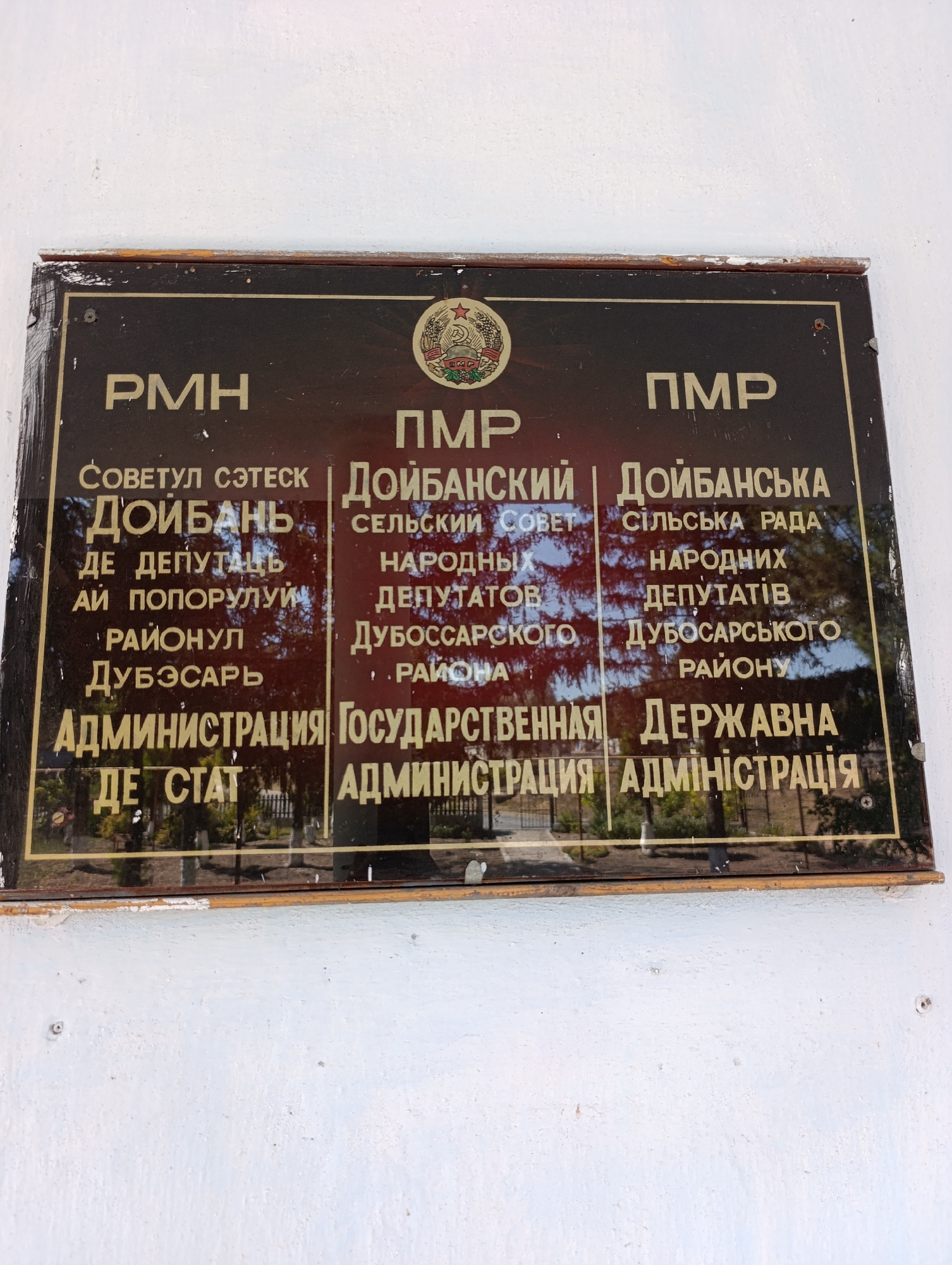 A close-up, eye-level, outdoor shot shows a dark, rectangular sign with gold lettering mounted on a white wall. The sign is bordered by a wooden frame. The sign displays text in multiple languages, including Russian and what appears to be Gagauz, as well as a Soviet-era emblem at the top center. The text refers to "PMH," "NMP," and various administrative terms such as "Sovety," "Deputatov," and "Administratsiya." It also includes place names like "Doibani" and "Dubossary." The reflection on the sign suggests it is a sunny day, with trees visible in the background. The sign appears to be old, with some wear and tear on its edges.
