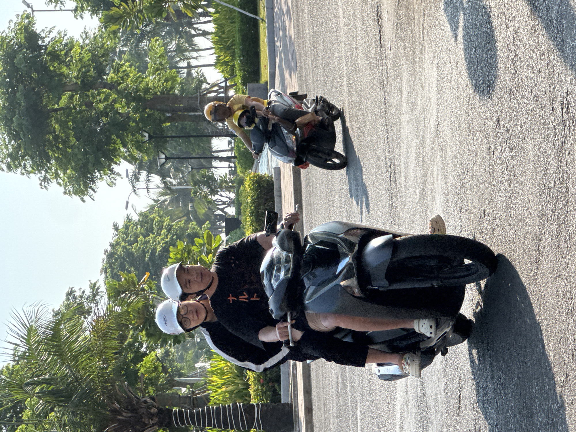 Two individuals are riding a dark-colored scooter on a paved road in Nha Trang, Vietnam. The person in the front, wearing a white helmet and glasses, is operating the scooter, while the person behind, also wearing a white helmet, is seated behind them. Both are dressed in casual clothing, with the driver wearing a black t-shirt with Japanese characters and the passenger wearing a black jacket with white stripes. Another scooter with two people, a man and a child, is visible further down the road, also traveling in the same direction. The environment suggests a tropical location, with palm trees and lush green foliage lining the road. The scene appears to be captured during daylight, with shadows cast on the asphalt, indicating the sun is present. The road is a multi-lane thoroughfare, and there are streetlights visible in the background. The overall impression is a common mode of transportation in Vietnam.