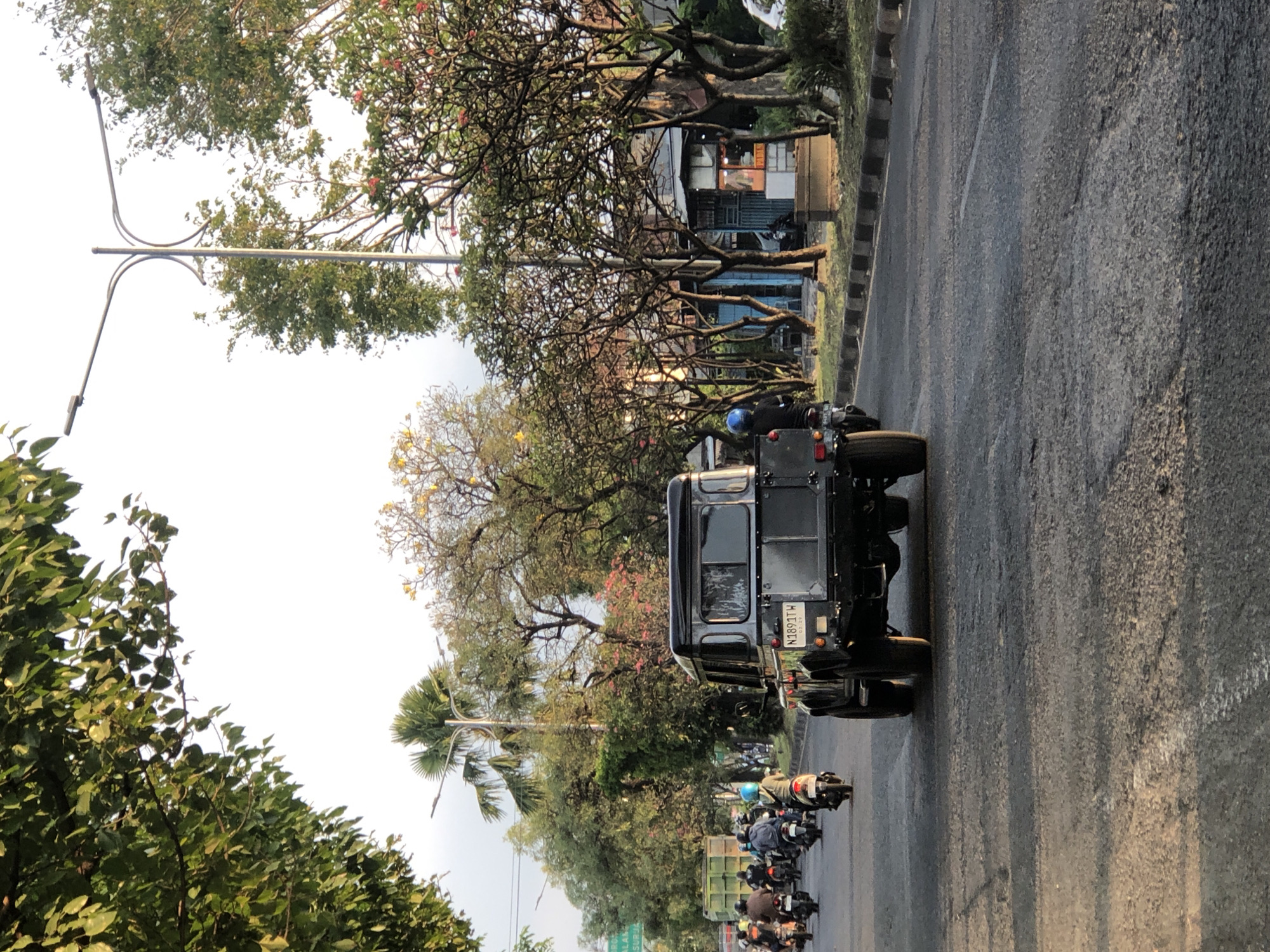 The image depicts a street scene in Gelam, Indonesia, during the day. A black SUV with the license plate N1891TW is driving away from the camera in the foreground. Behind the SUV, a group of motorcycles and their riders are also traveling in the same direction. The road is paved and shows signs of wear. On the sides of the road, there are numerous trees, some with green foliage and others with pink and yellow flowers. A street light with two lamps is visible in the middle ground. In the background, there are buildings and more trees. The sky is bright, indicating daylight.