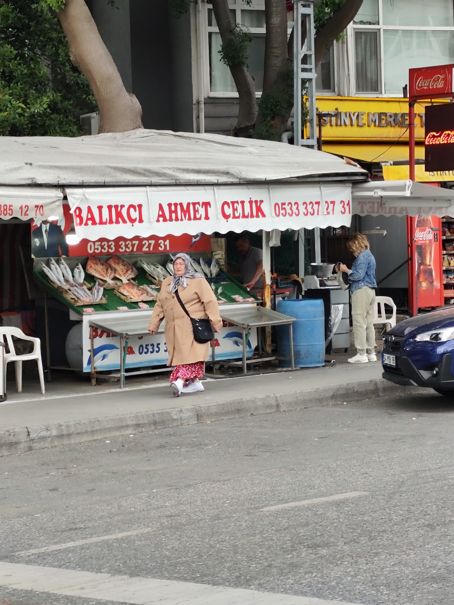 The image depicts a street scene in Sarıyer, Turkey. A fishmonger's shop, "BALIKÇI AHMET ÇELİK," stands at the edge of the sidewalk with a sign displaying phone numbers. A display of fish is visible under the canopy. An elderly woman wearing a headscarf, a beige coat, and a patterned red dress is walking in front of the shop. A customer in a denim jacket is by a scale, possibly purchasing fish, while a man appears to be working at the shop in the background. A blue car is parked near the shop, and a Coca-Cola vending machine stands adjacent to the fish shop. The background includes buildings and trees. A sign for "İSTİNYE MERKEZİ" (İstinye Center) is visible above the fish shop.