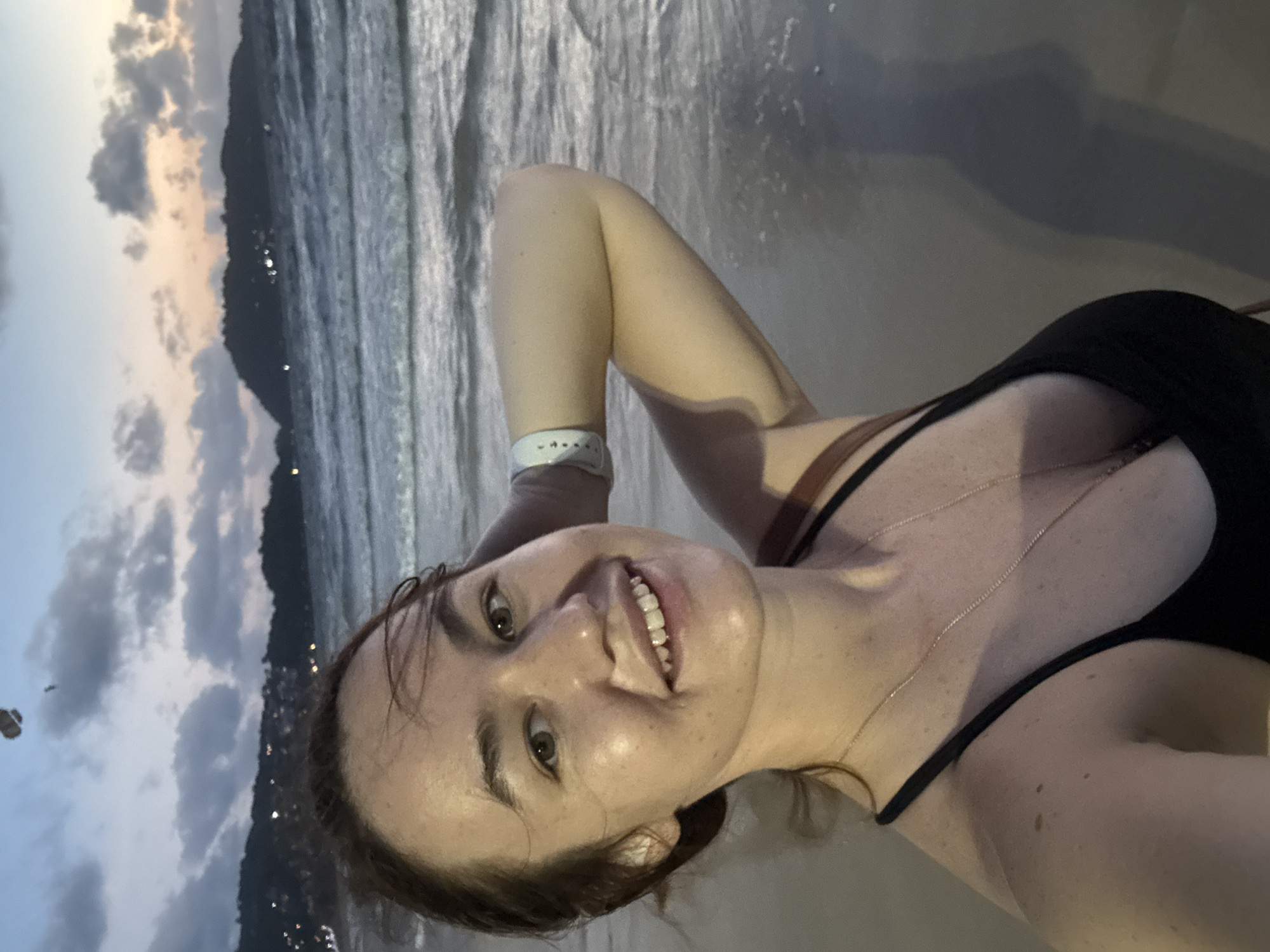 A woman with light skin and reddish-brown hair is taking a selfie on a beach in Patong, Thailand, at dusk. She is wearing a black bikini top and a thin gold necklace. Her right arm is raised and bent, with her hand behind her head. She is smiling and looking directly at the camera. Her left arm is not fully visible. The ocean is in the background, with gentle waves rolling onto the sandy shore. In the distance, hills with lights are visible along the coastline, indicating the city of Patong. The sky is a mix of blue, grey, and orange, suggesting the sun has recently set. A paraglider can be seen as a dark silhouette against the sky in the upper left portion of the image.