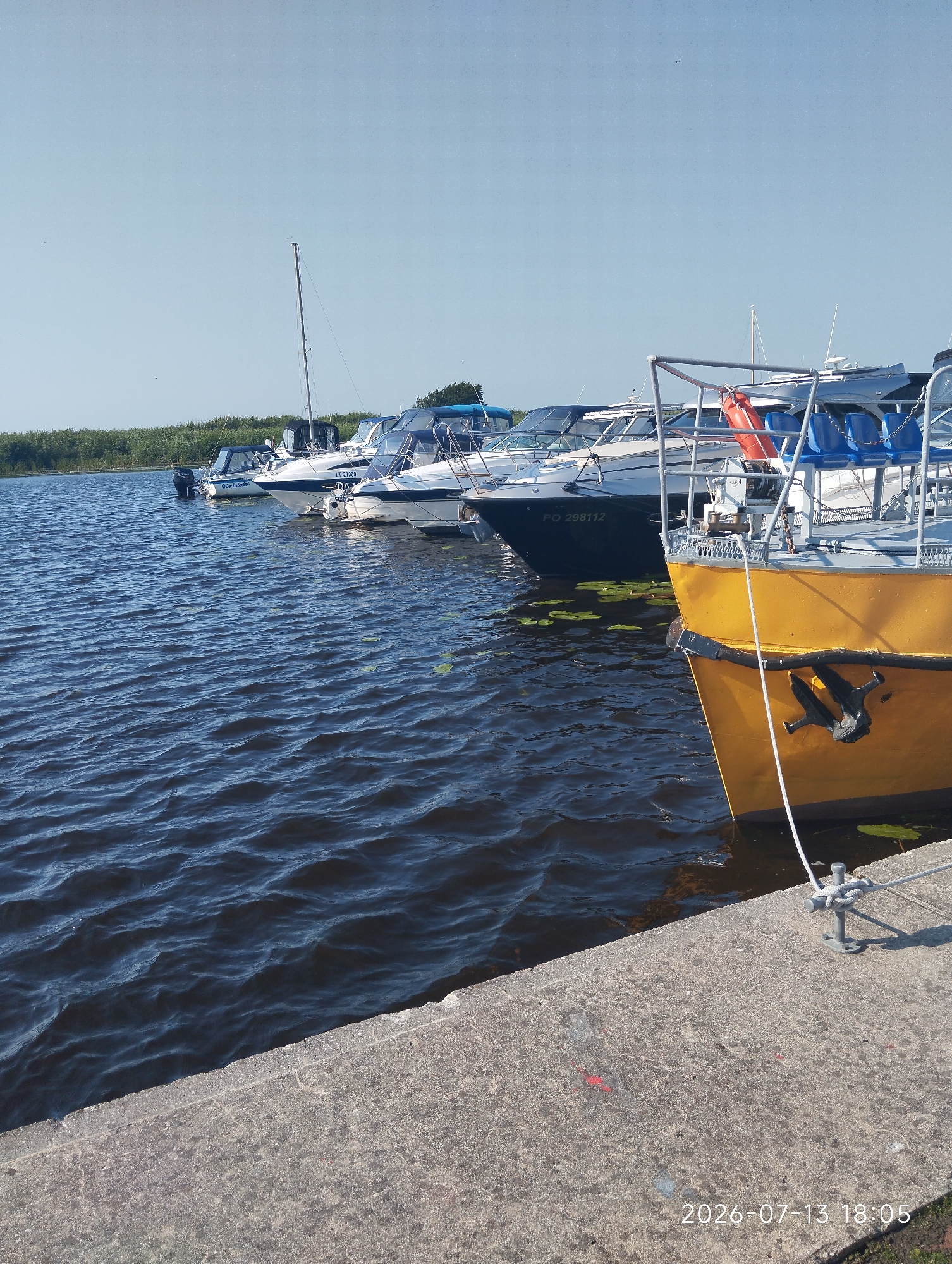 The image shows a sunny day at a marina in Dreverna, Lithuania, with several boats docked. In the foreground, a yellow boat is moored to a concrete pier, with its anchor visible. The water is dark blue and choppy, with some lily pads floating on the surface. In the background, a line of white and blue-capped boats are docked along the pier. The sky is clear and blue, with a few distant birds. A line of green vegetation runs along the far bank of the river. The timestamp on the image reads 2026-07-13 18:05.