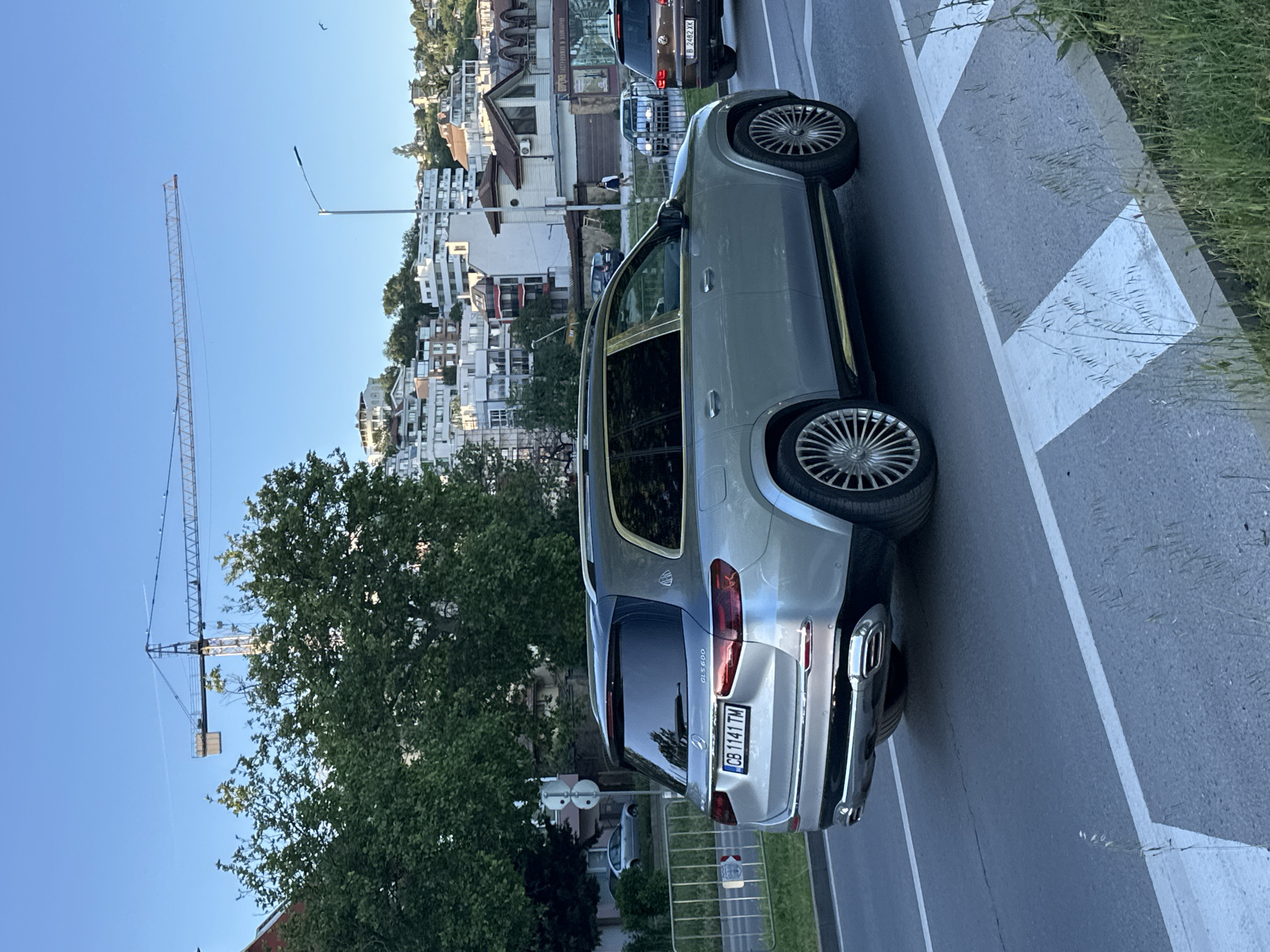 The image shows a silver Mercedes-Maybach GLS 600 parked on a street in Varna, Bulgaria. The car's license plate reads "CB 1141 TM." Behind the Maybach, a brown Volkswagen SUV is partially visible. In the background, there are buildings, including residential structures, indicating an urban environment. A construction crane is visible to the left, suggesting ongoing building activity in the area. The street has painted white markings, indicating a crosswalk or pedestrian zone. The overall setting is urban with a mix of residential and commercial areas. Vegetation, likely trees and grass, is visible on the right side of the frame.