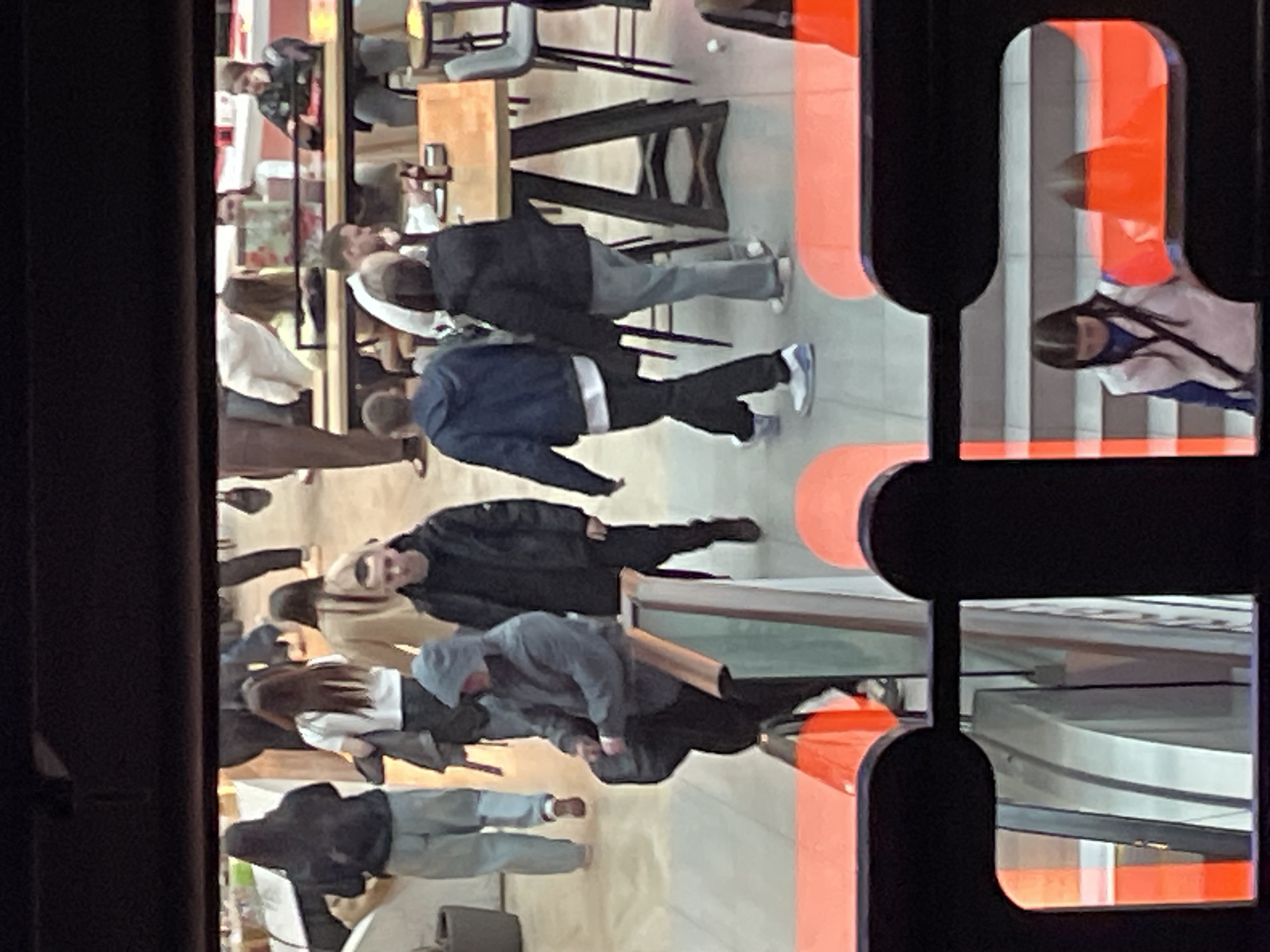 The image depicts a partial, distorted view of an interior space with multiple people. The location appears to be in Saint Petersburg, Russia, based on the architecture of the interior.
The scene includes several individuals walking and standing. Most are dressed in casual clothing.
Visible objects include tables, chairs, and glass surfaces, indicating a cafe, restaurant, or similar public space. The perspective is from behind a dark, abstractly designed structure with curved, rectangular cutouts, casting the scene as if viewed through an artistic frame. The lighting is bright, possibly artificial, highlighting the interior scene.