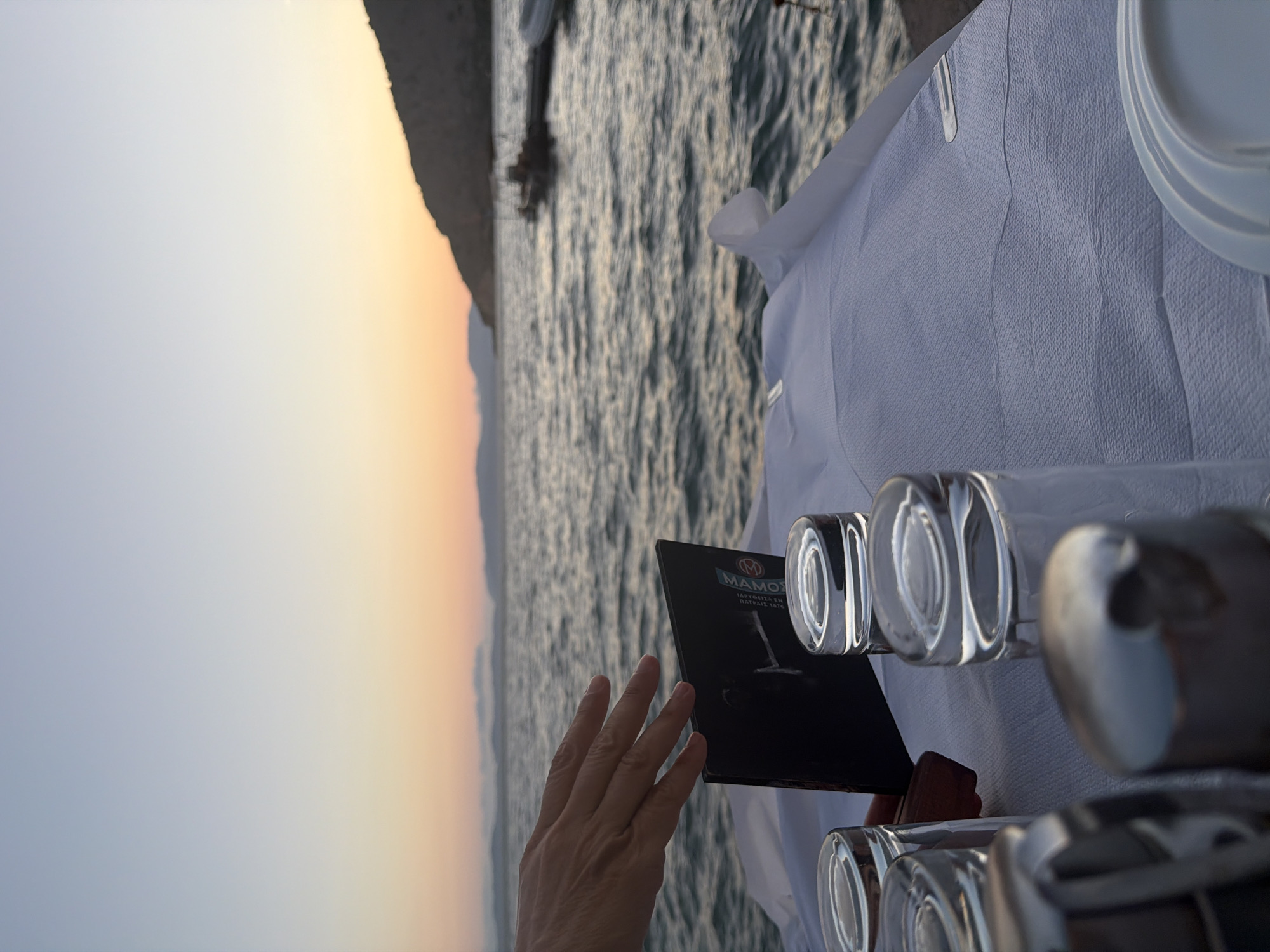 An outdoor restaurant table is set for a meal by the sea at dusk in the Municipal Unit of Alyzia, Greece. A hand, likely belonging to a patron or server, is reaching out towards a dark-colored menu. Several clear glasses are placed on a white tablecloth, and a stack of white plates is visible on the right side of the frame. The sea, with moderate ripples, extends to the horizon, where a rocky island is silhouetted against a gradient sky of pale blue and soft orange hues, indicative of sunset. A few boats are moored in the distance. The overall impression is one of a serene evening dining experience by the water.