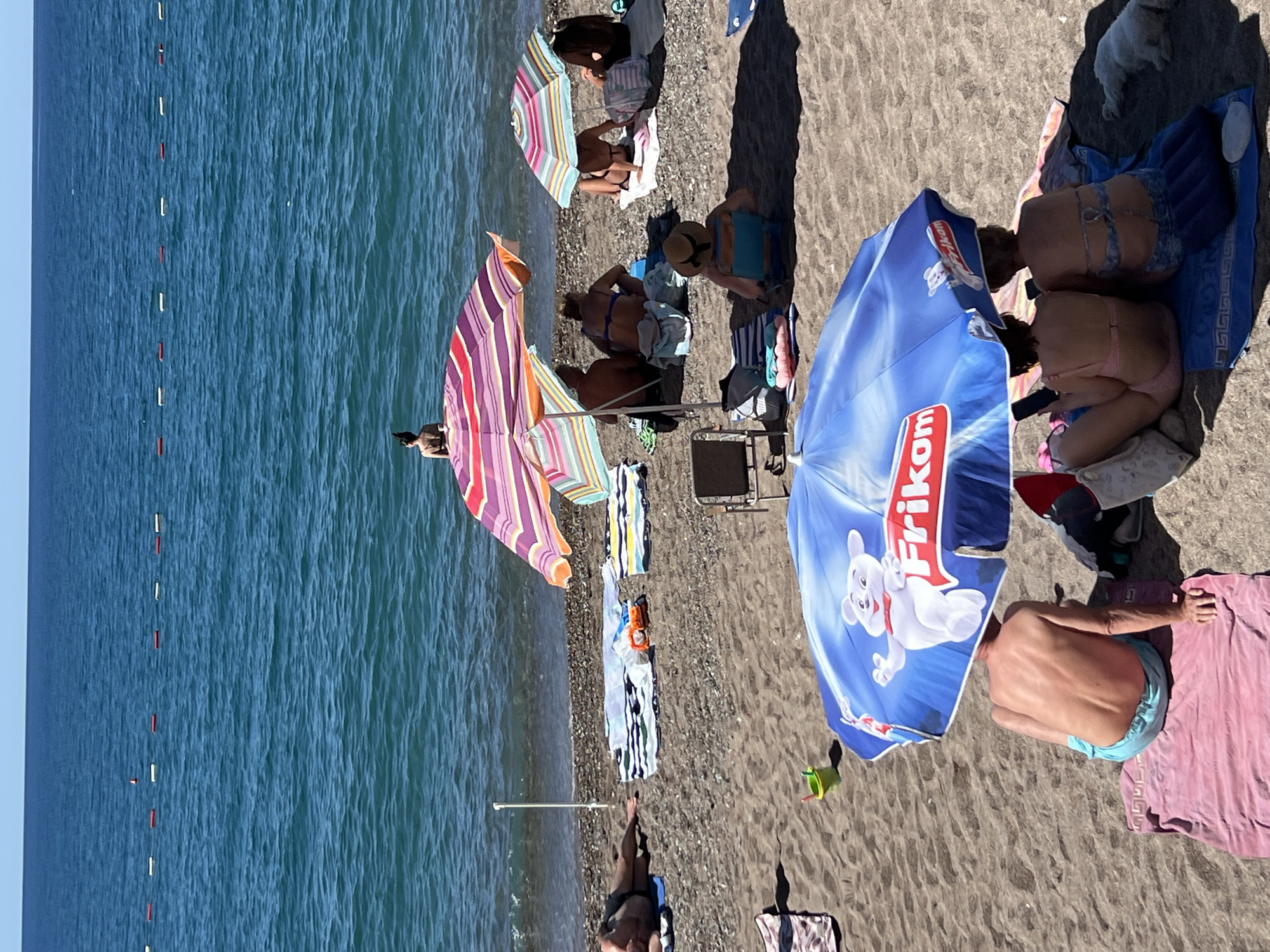 The image depicts a sunny day at the beach in Rafailovići, Montenegro. The sea is calm and blue, with a line of buoys marking a swimming area. On the sandy and pebbly beach, several groups of people are relaxing under colorful umbrellas. In the foreground, two women are seated under a large blue umbrella with a "Frikom" logo, looking at their phones. To their right, a small white dog is lying down. Behind them, a man in light blue swimming trunks sits on a pink towel. Further back, more people are visible under striped umbrellas. One person is lying on a towel in the sand to the left. In the distance, a lone figure stands near the water's edge. Beach chairs and towels are scattered across the scene, indicating a leisurely day at the resort.