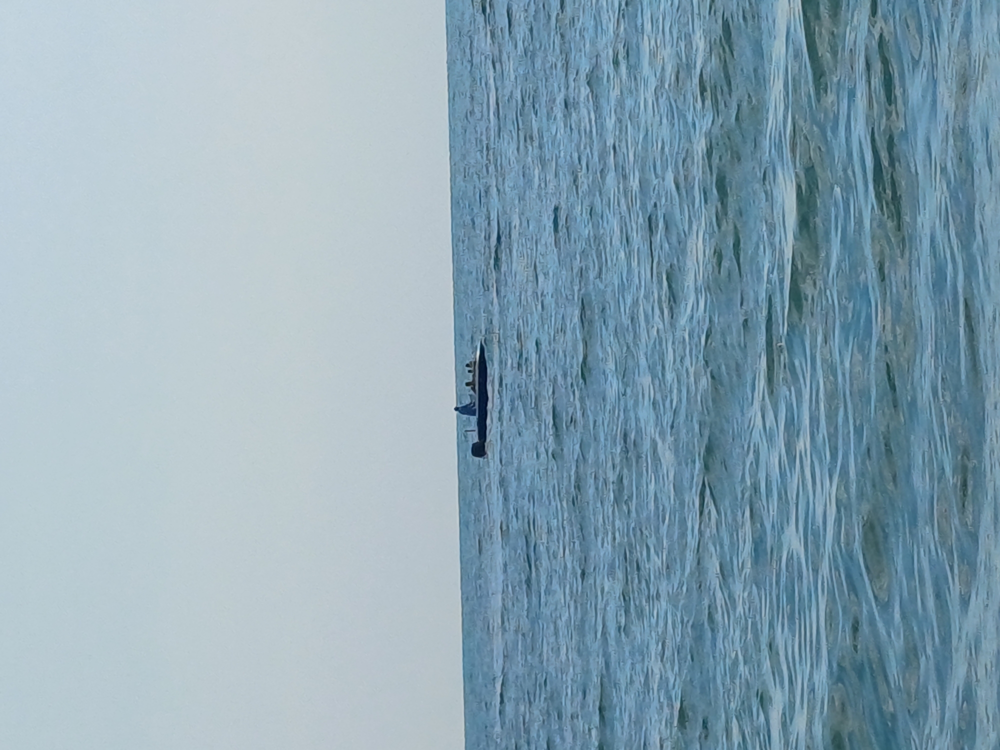 A small, dark-colored motorboat with at least three individuals on board is navigating on the tranquil, rippling blue waters of the Black Sea, with a faint, light blue sky above. The boat, positioned on the left side of the image, appears to be moving away from the viewer, with one person standing at the stern, possibly operating the vessel. The horizon line between the sea and sky is clearly visible. The overall scene is serene and depicts a calm day at sea. The location is the coast of Sochi, Russia.