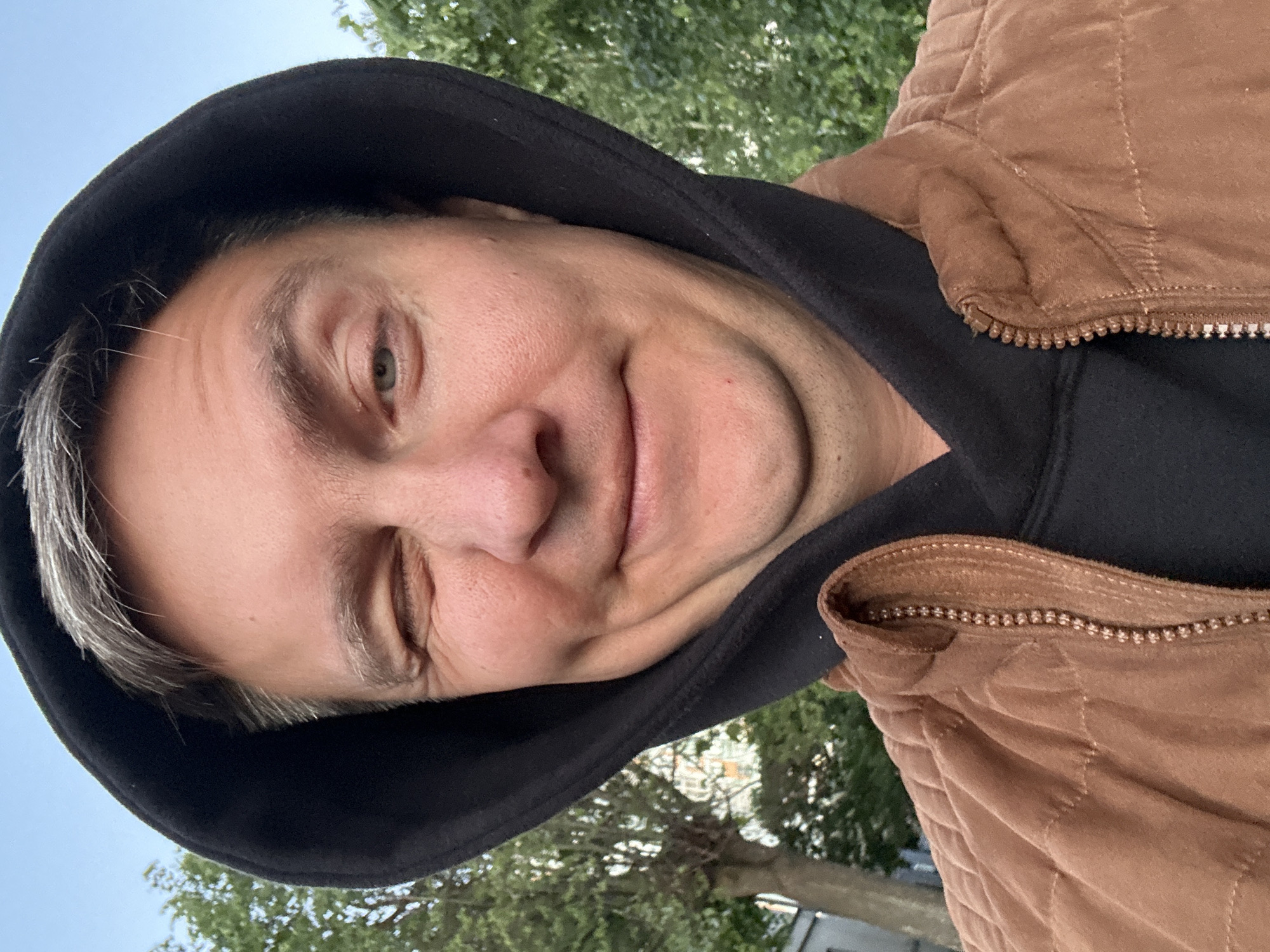 A selfie of a middle-aged man with fair skin, wearing a black hooded sweatshirt and a brown quilted jacket, is rotated approximately 90 degrees counterclockwise. He is winking with his left eye and smiling slightly. He has short, graying hair. The background includes trees and a building. The location is not discernible as Minsk, Belarus, from the visual information provided.